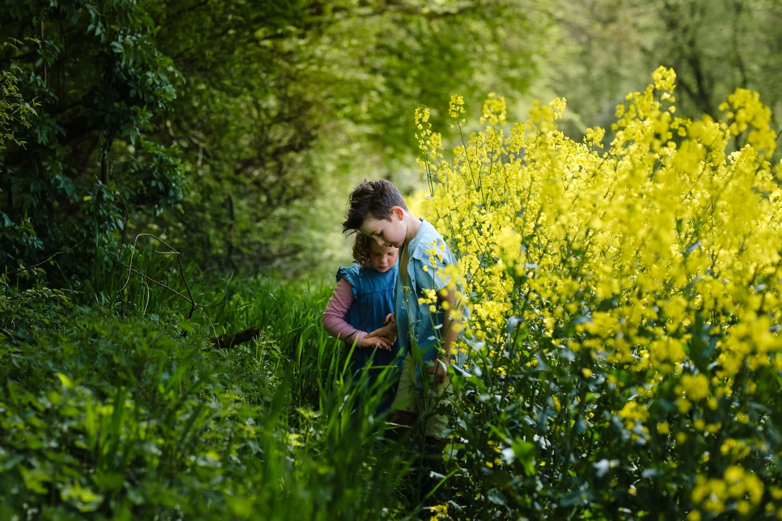 Two children, a boy and a girl, are exploring plants in a lush green outdoor setting with yellow flowers in bloom.