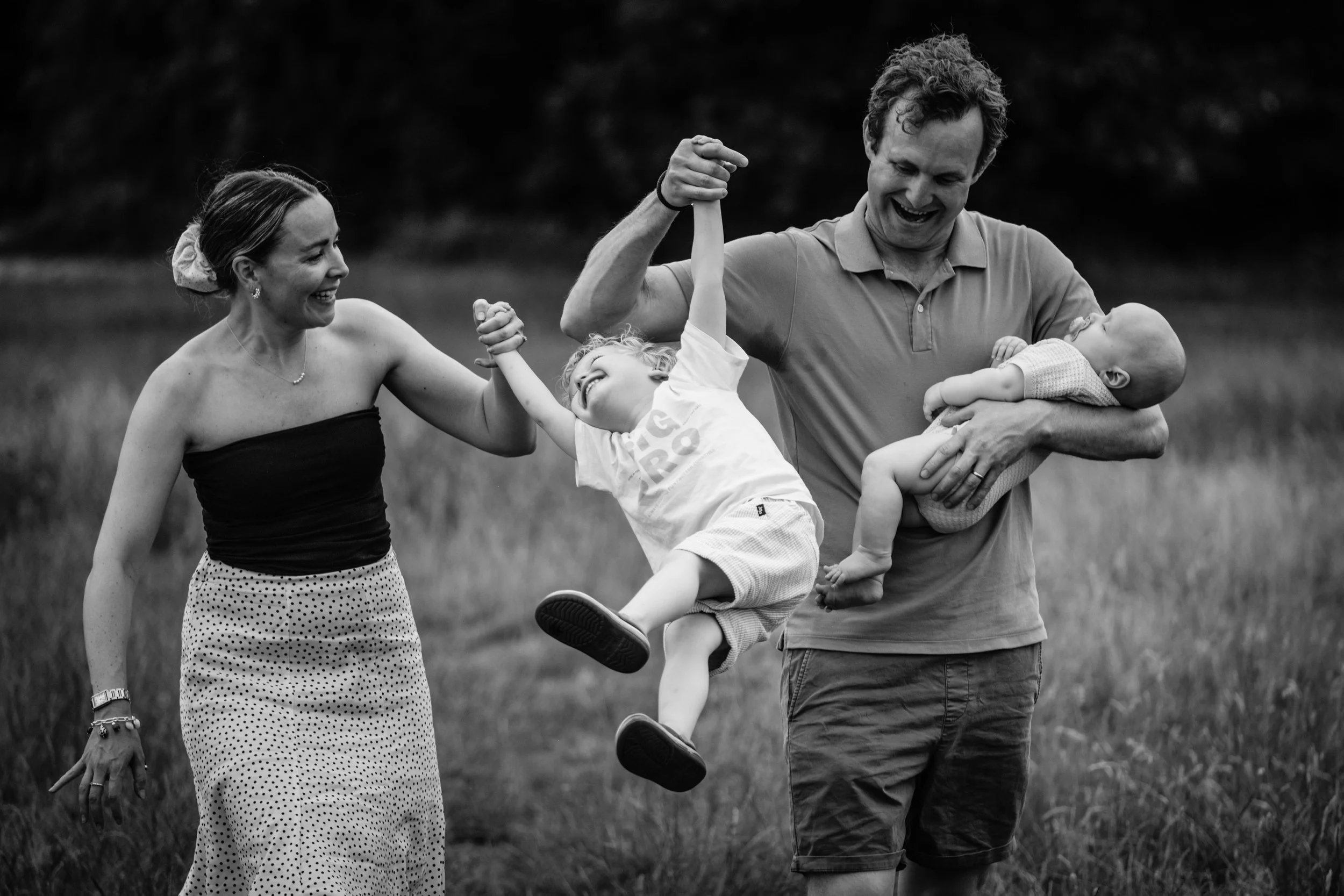 A family of four enjoying a playful moment outdoors, with a woman in a strapless top and polka dot skirt, a man holding a young boy and a baby, all smiling and appearing happy.
