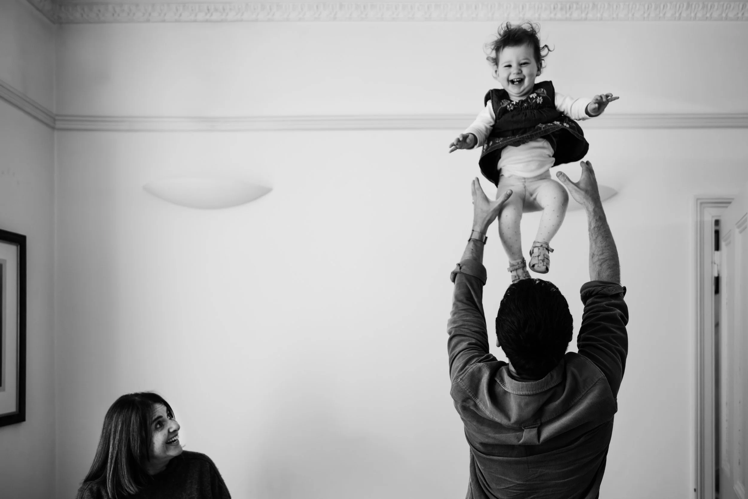 A man lifts a smiling child into the air while a girl looks up at them and laughs.