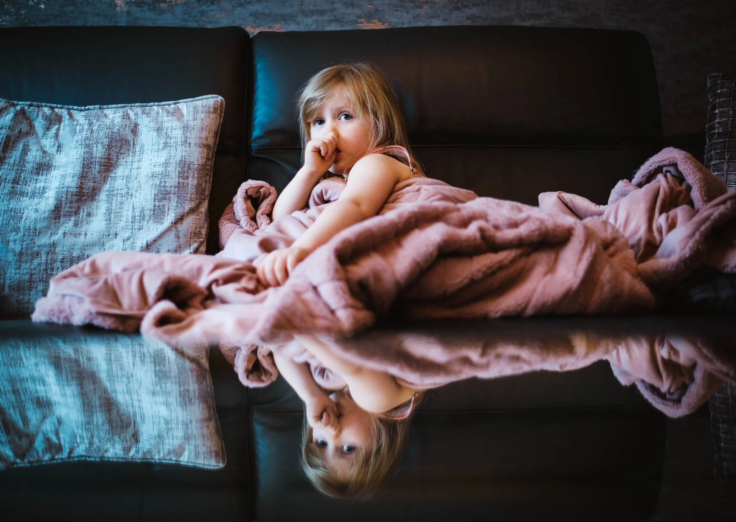 A young girl with blonde hair and blue eyes lying on a black leather couch, wearing a pink sleeveless dress, with a pink blanket covering her and a pillow nearby, looking at the camera. The image show her reflection in a glass table top.