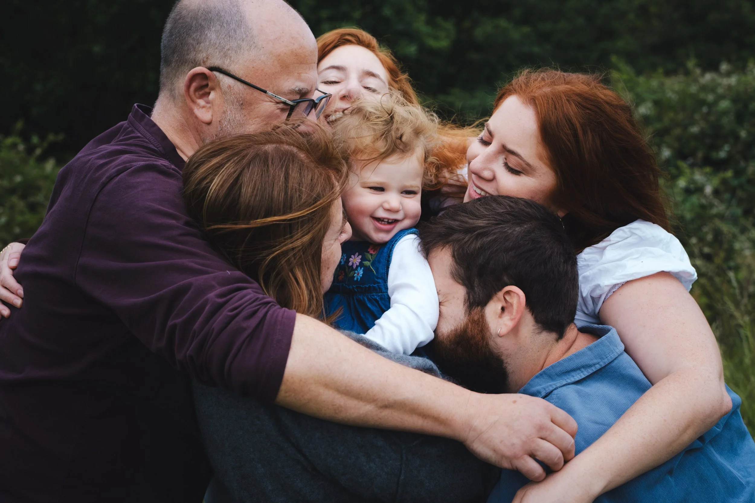 A large group of people, including children and adults, hugging tightly outdoors in a joyful group hug with green trees in the background.
