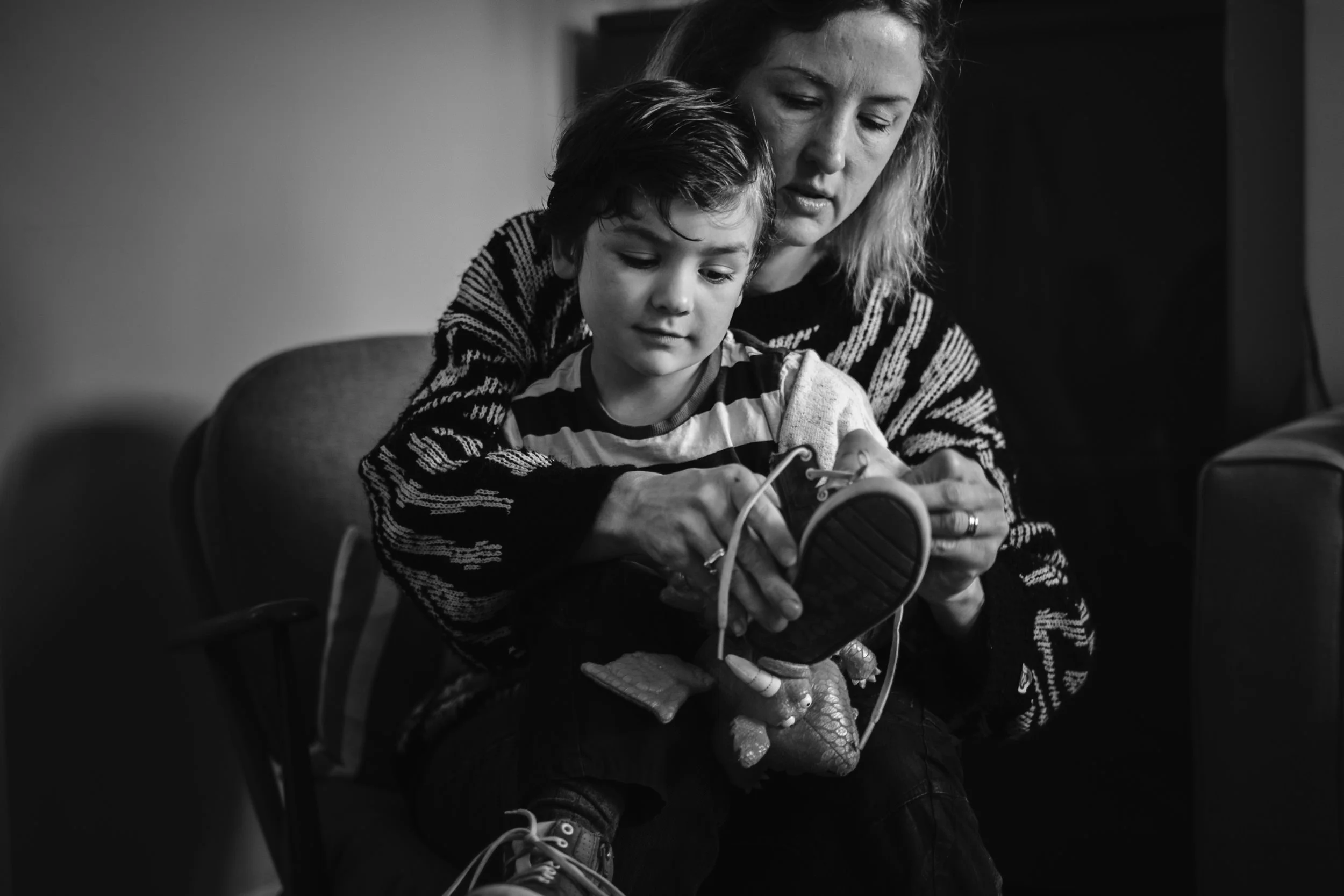 A woman and a young boy sitting closely together, looking at a smartphone. The woman is helping the boy with the device in a caring manner. The image is in black and white.