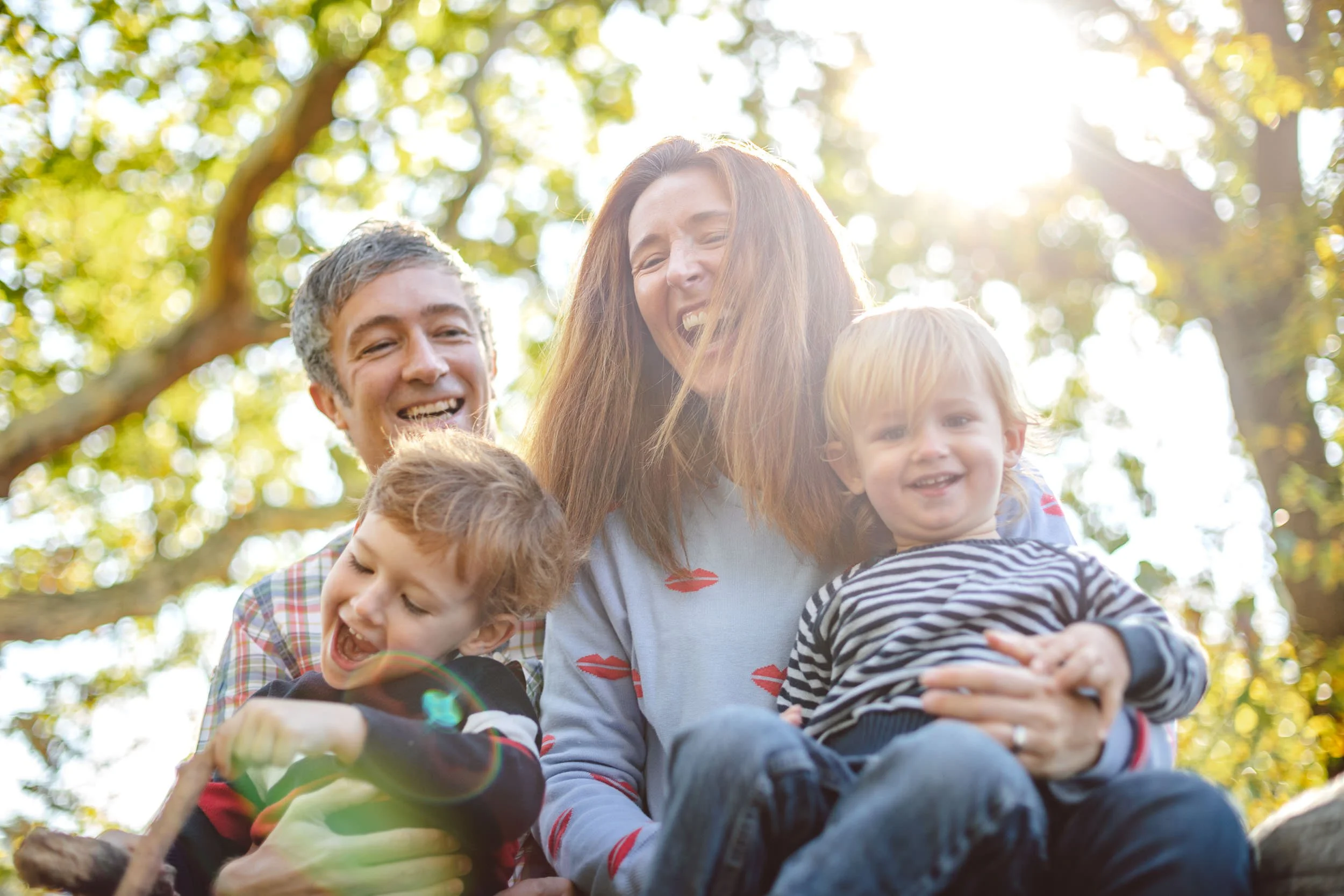 Group of happy children and a woman outdoors under trees, smiling and laughing, with sunlight shining through the foliage.