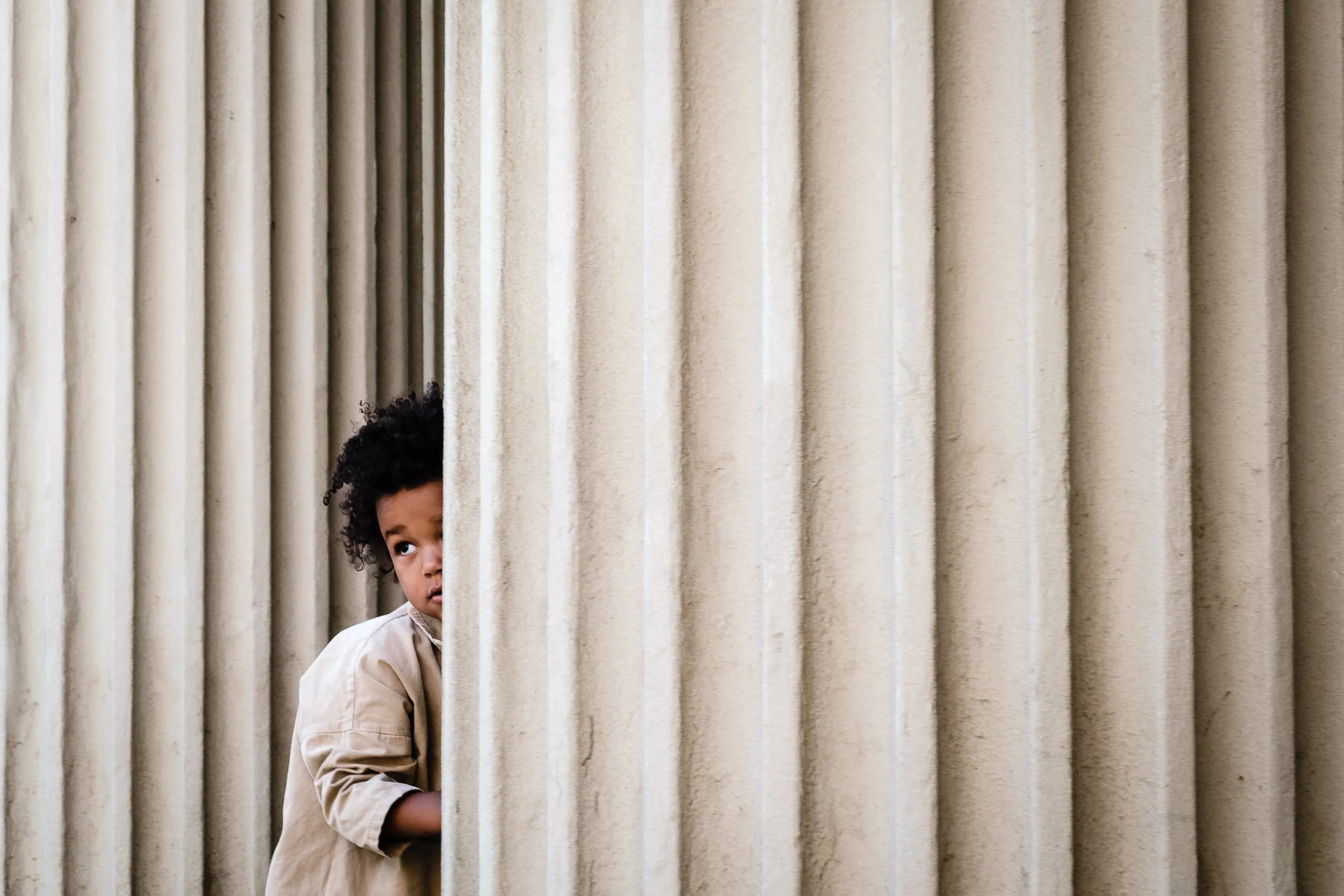 A young girl with curly hair peeking from behind vertical beige stone or concrete columns.