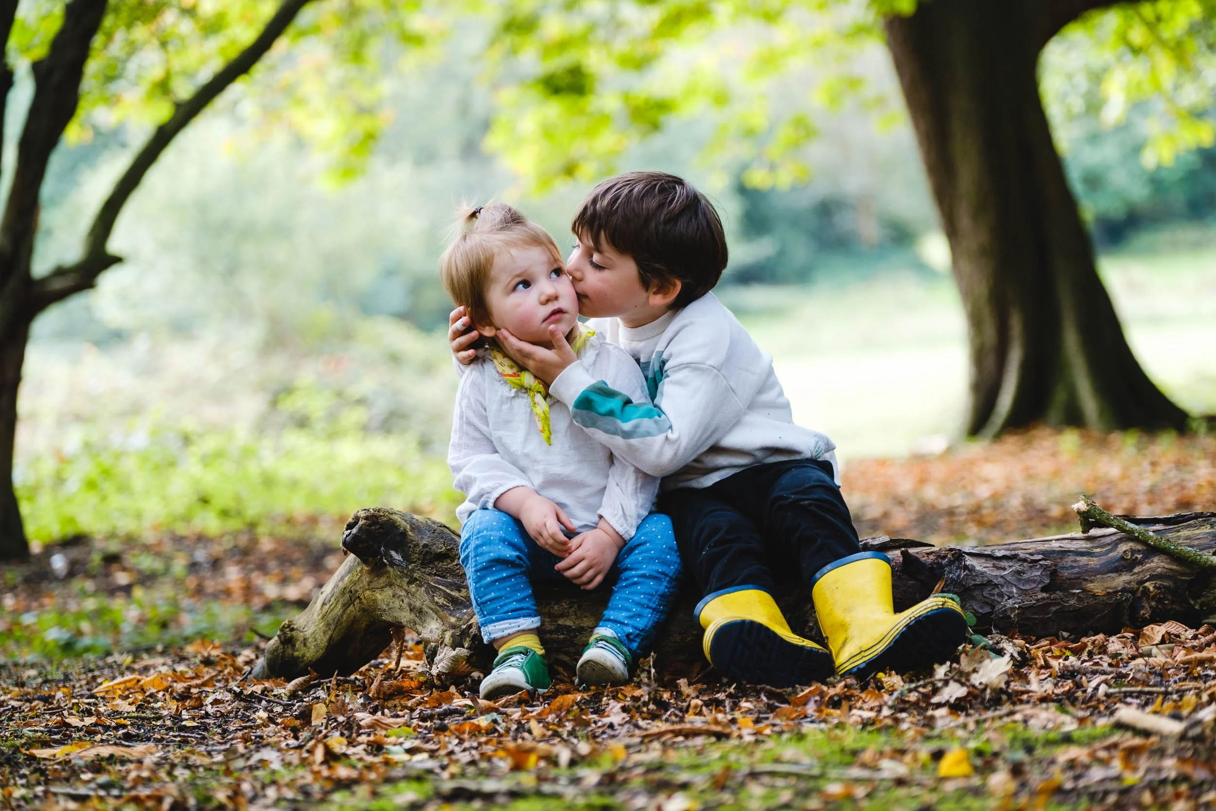 A boy and a girl sitting on a fallen log in a forest, with the boy giving the girl a kiss on the cheek. The girl looks surprised, and there are autumn leaves on the ground. The background shows green trees and foliage.