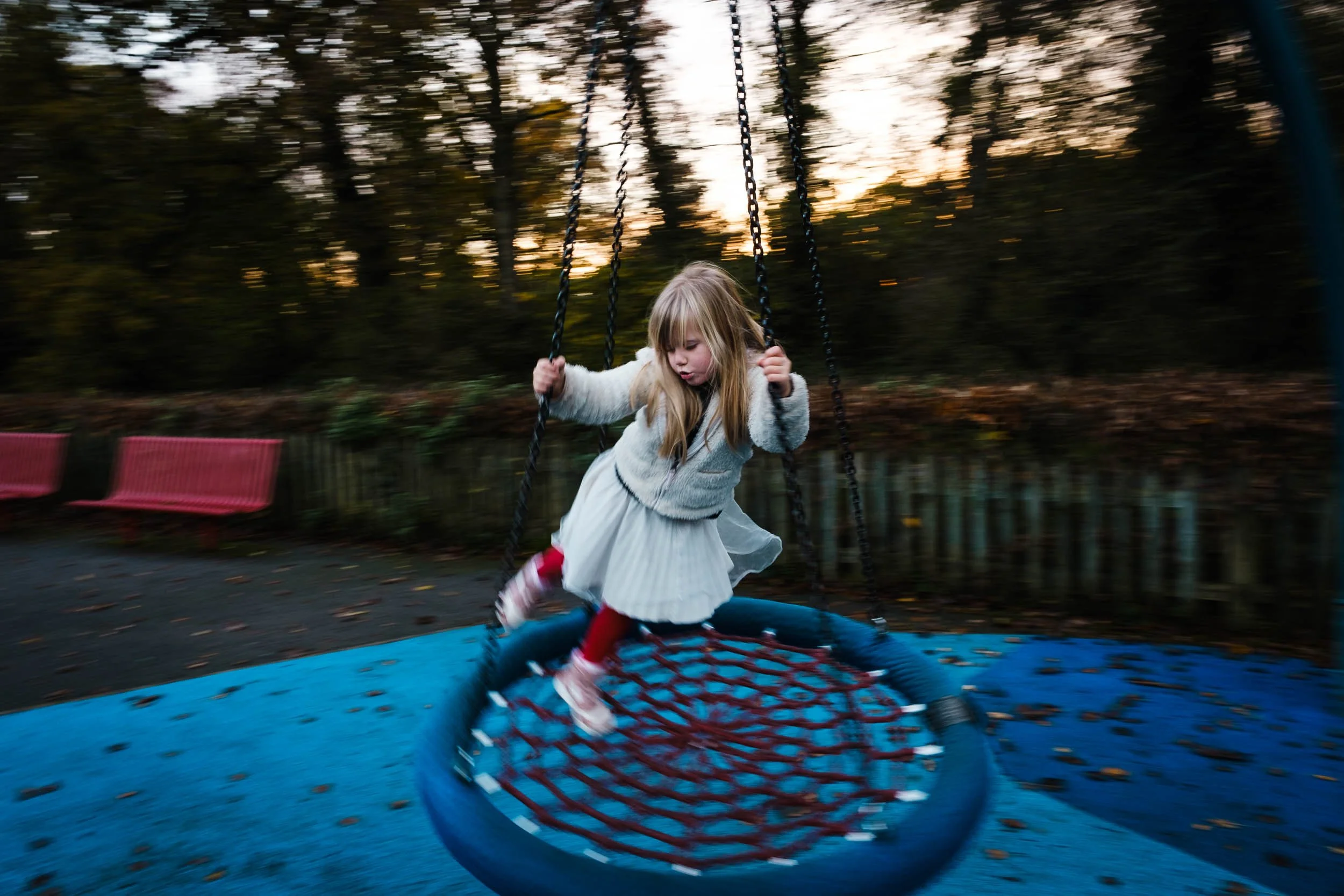 A young girl with blonde hair, wearing a white sweater and red shoes, swinging on a blue playground swing set during sunset in a park with trees and a red bench visible in the background.