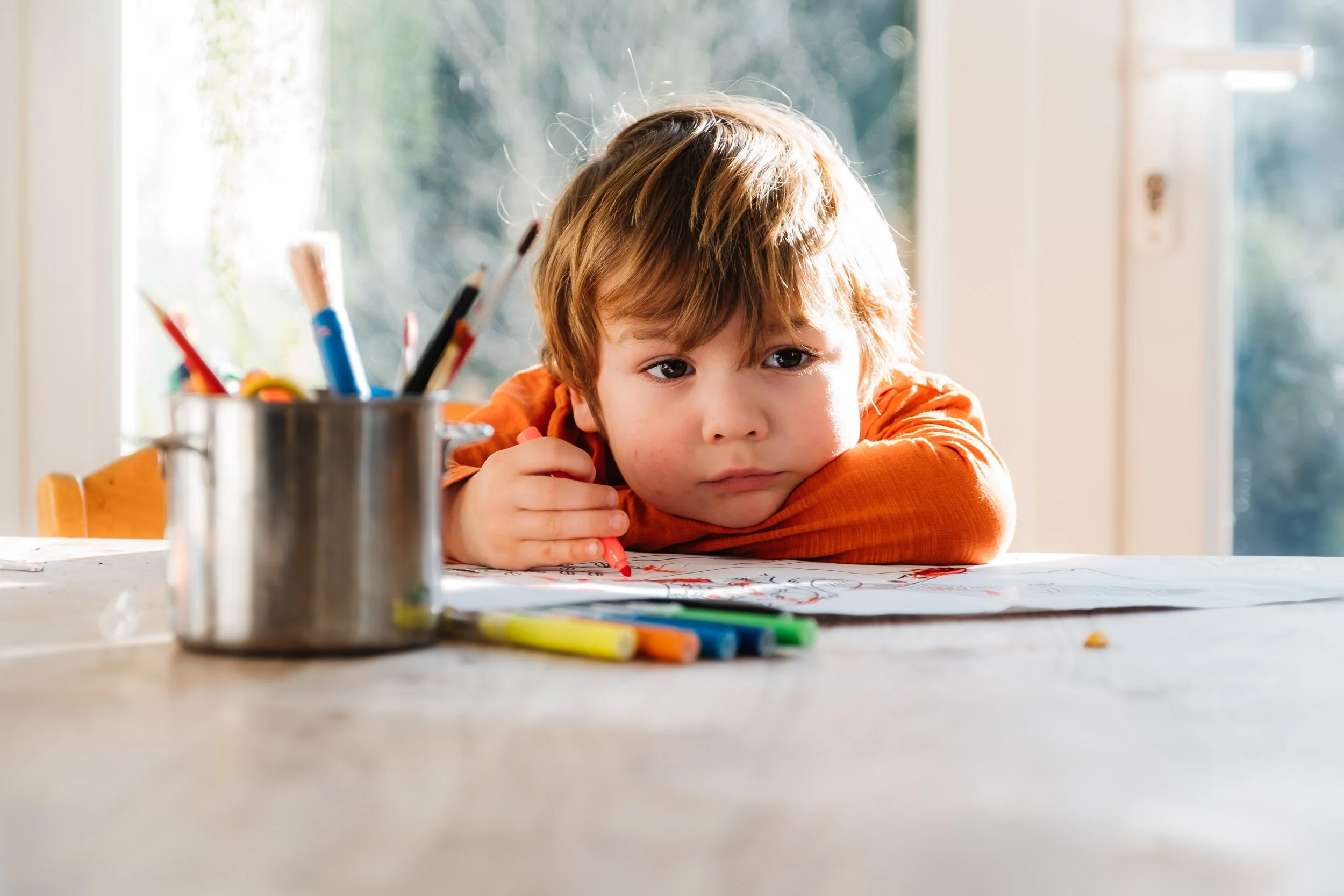 A young boy with light skin and brown hair drawing with a red crayon at a table in front of a window.