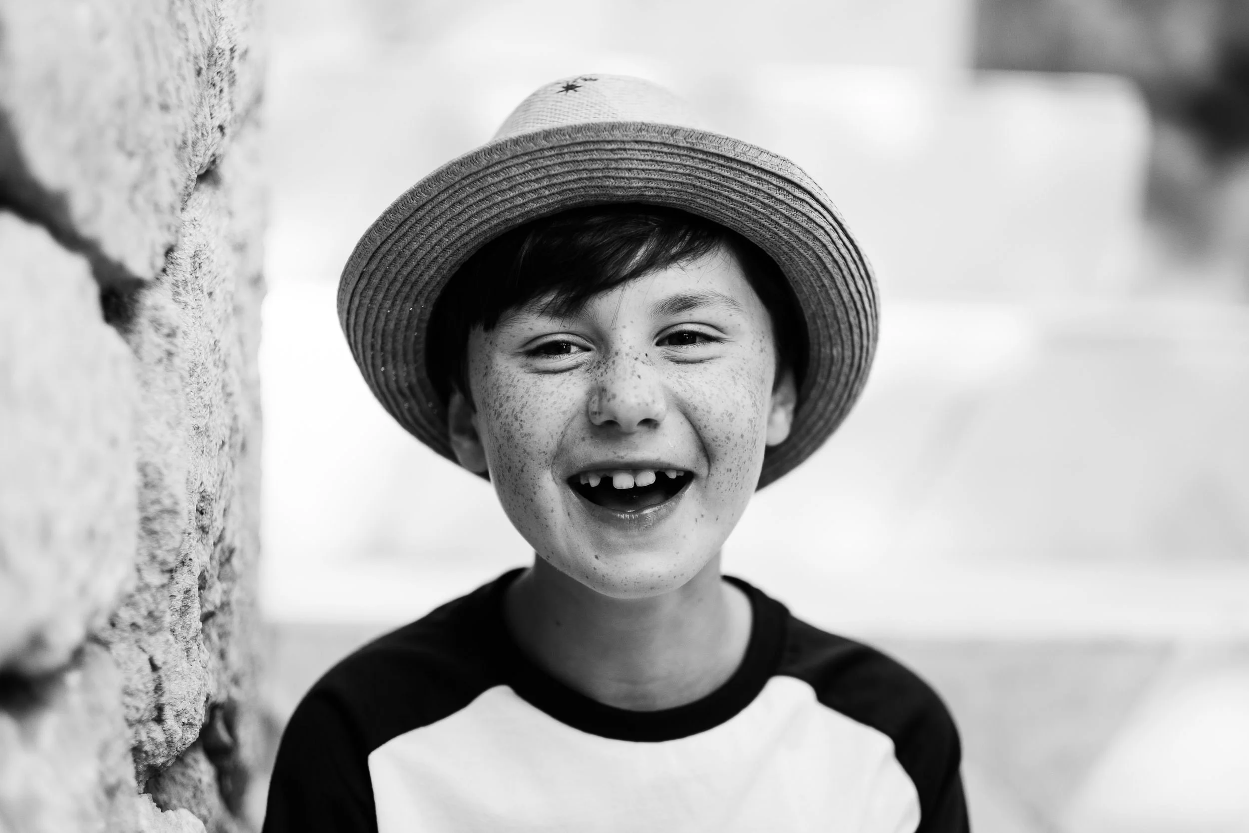 A smiling boy with freckles wearing a wide-brimmed straw hat and a black and white striped shirt, standing next to a textured brick wall outdoors.