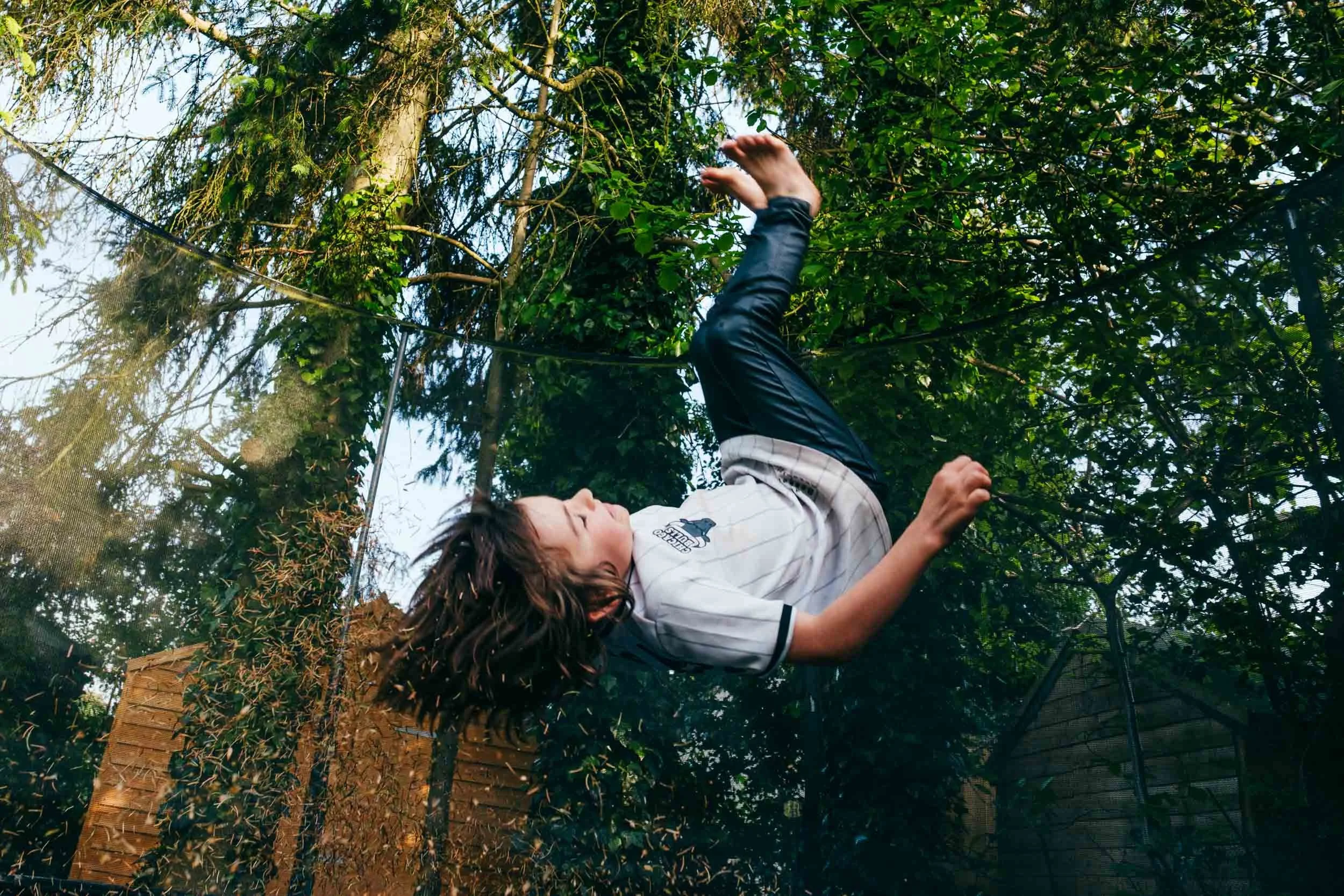A young boy upside down, bouncing on a trampoline, surrounded by green trees and foliage.