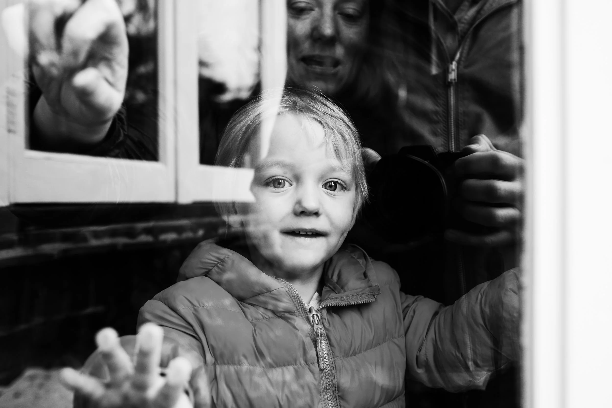 A young boy with light hair and eyes, wearing a puffer jacket, looks through a glass window outdoors. His mother stands behind him, also looking through the window. The photo is in black and white.