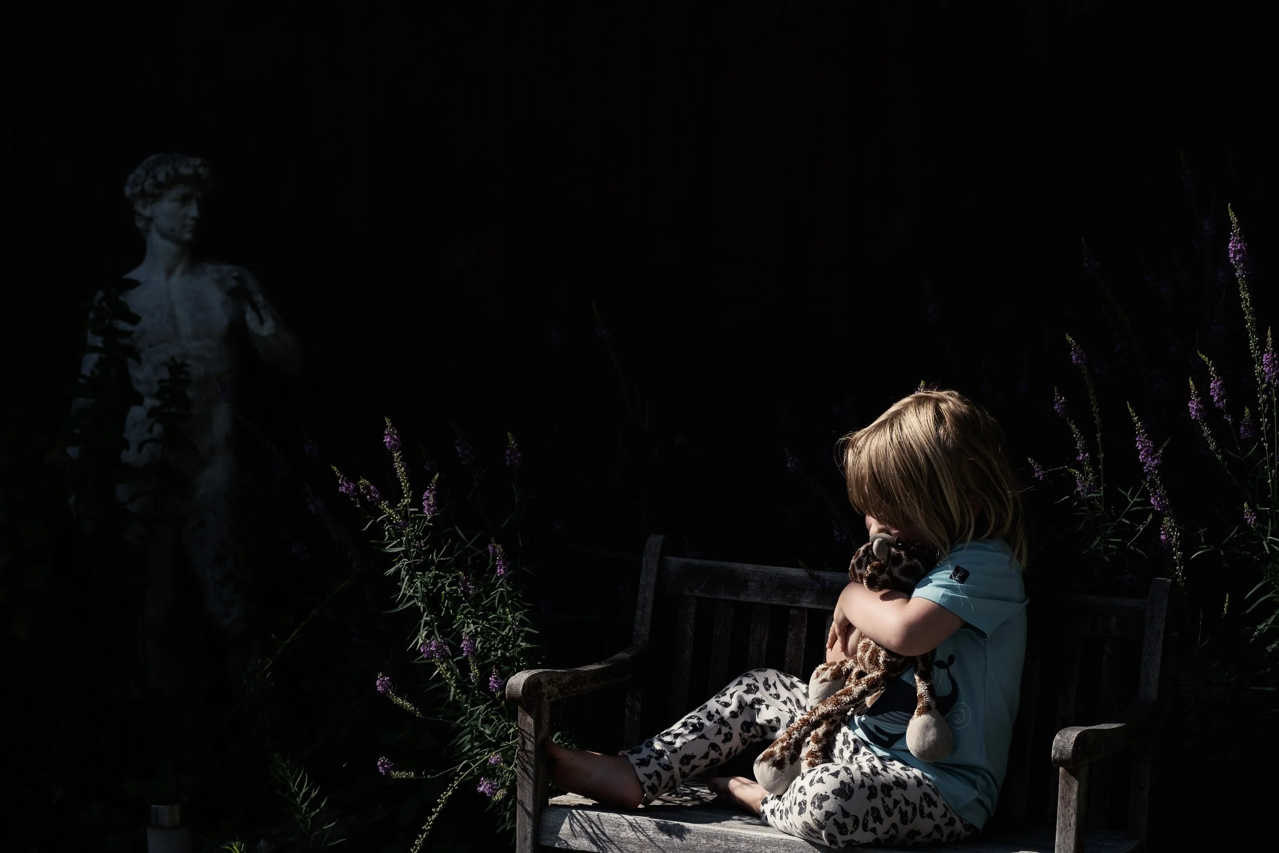 A young girl sitting on a wooden bench with her eyes closed, holding a stuffed animal, in a dark outdoor setting with purple flowers and a stone statue of a woman in the background.