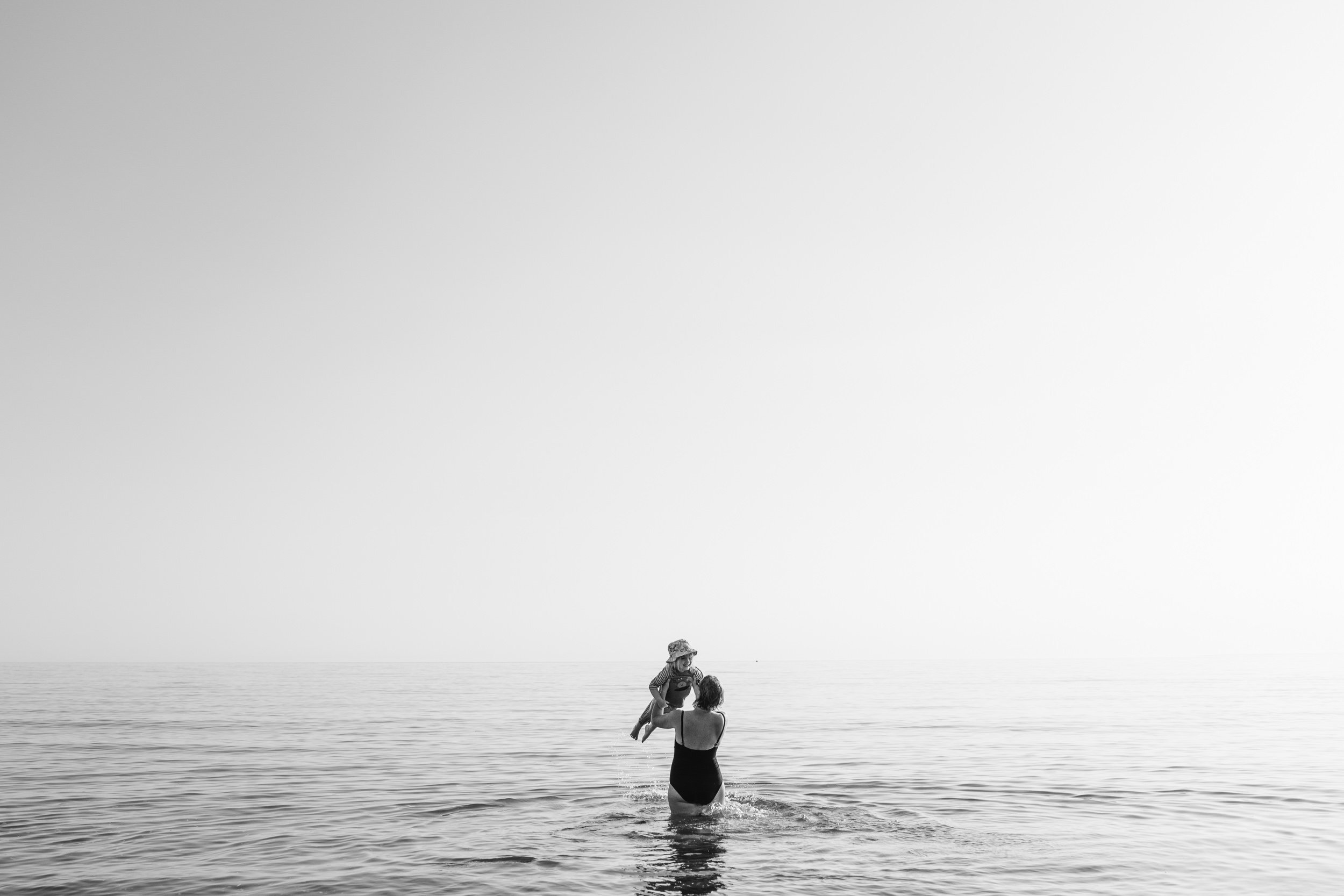 A woman in a swimsuit holding a young girl in a striped shirt and hat at the beach near the water's edge, with the ocean and sky in the background.