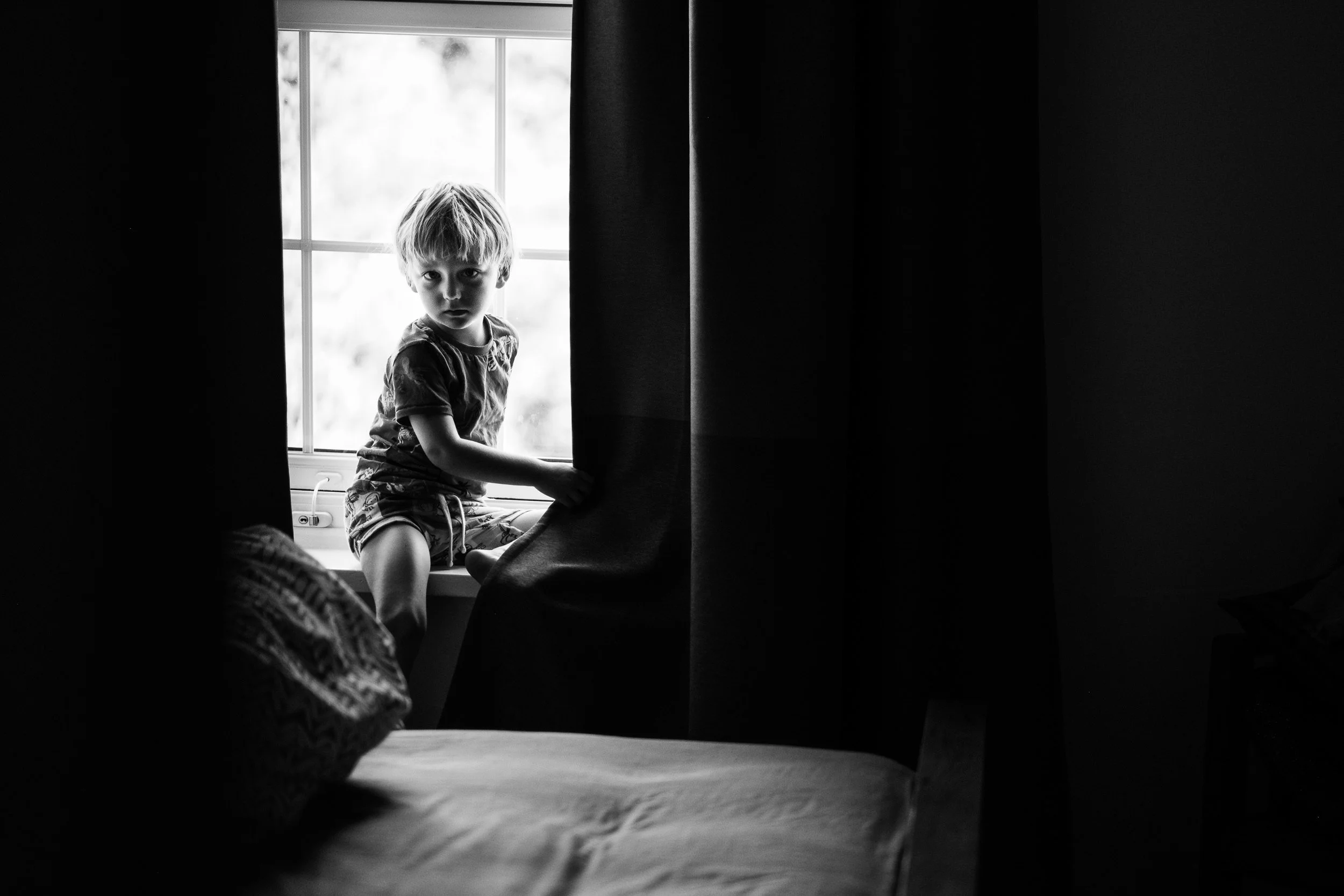 A young boy with blonde hair sitting on a window ledge, looking directly at the camera. The photo is in black and white, with dark curtains partially covering the view of the boy. The background visible through the window is bright, contrasting with 