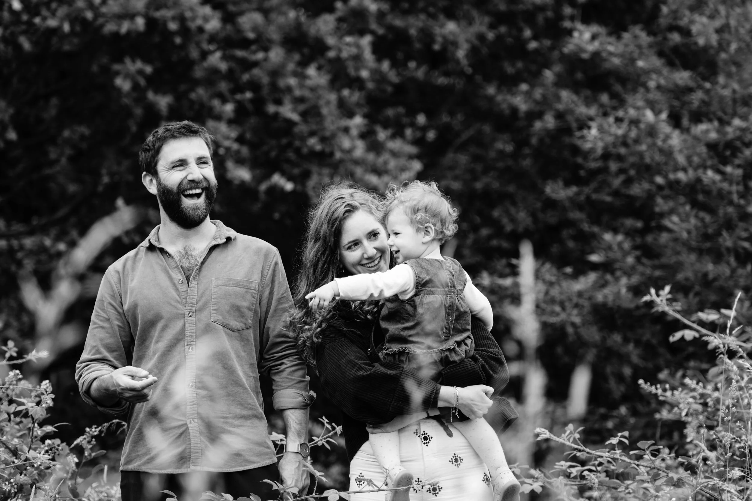 A happy family outdoors, with a bearded man and a woman holding a young girl, all smiling and enjoying a moment together.