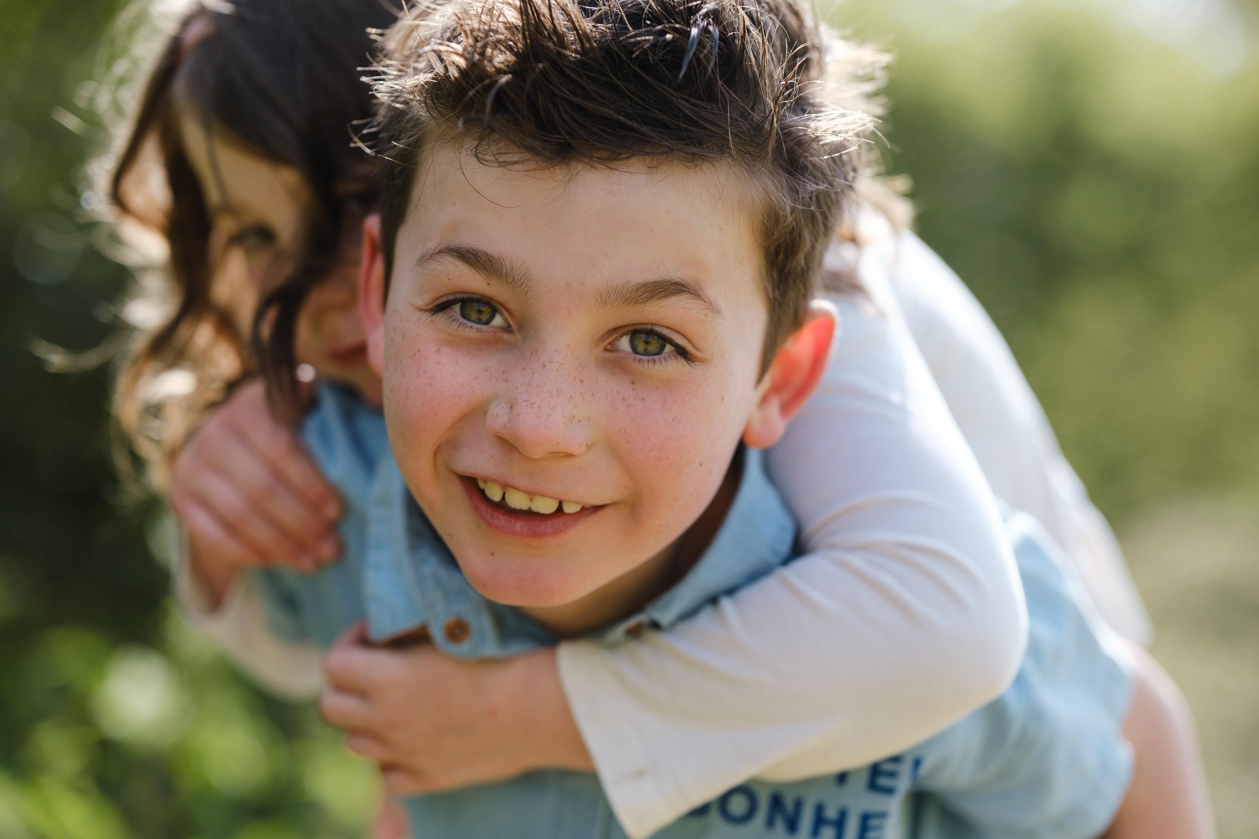 Close-up of a smiling boy with green eyes and freckles, leaning forward outdoors with blurred greenery in the background.