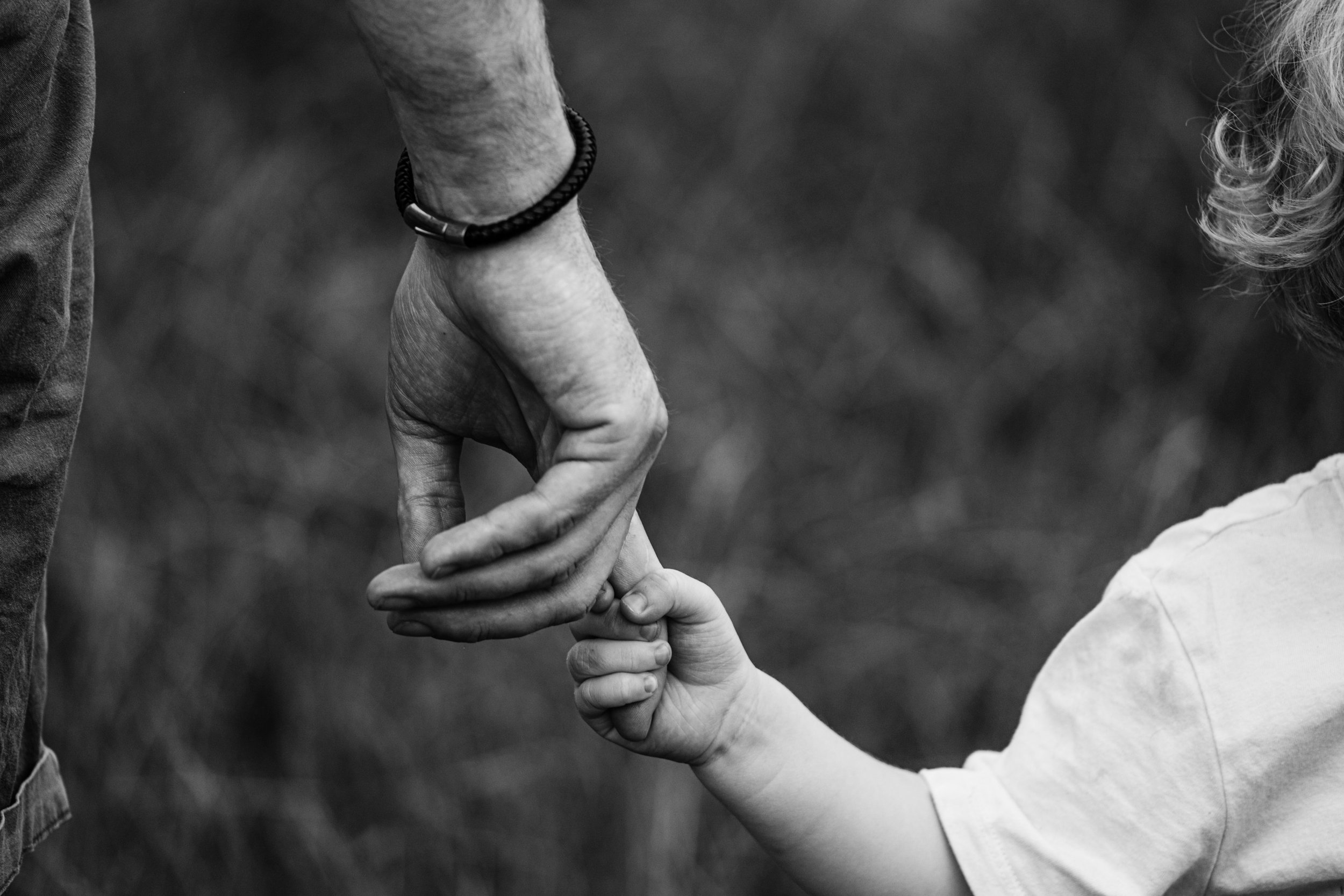 A black and white photo of an adult hand holding a child's hand, with the adult wearing a bracelet and the background blurred.