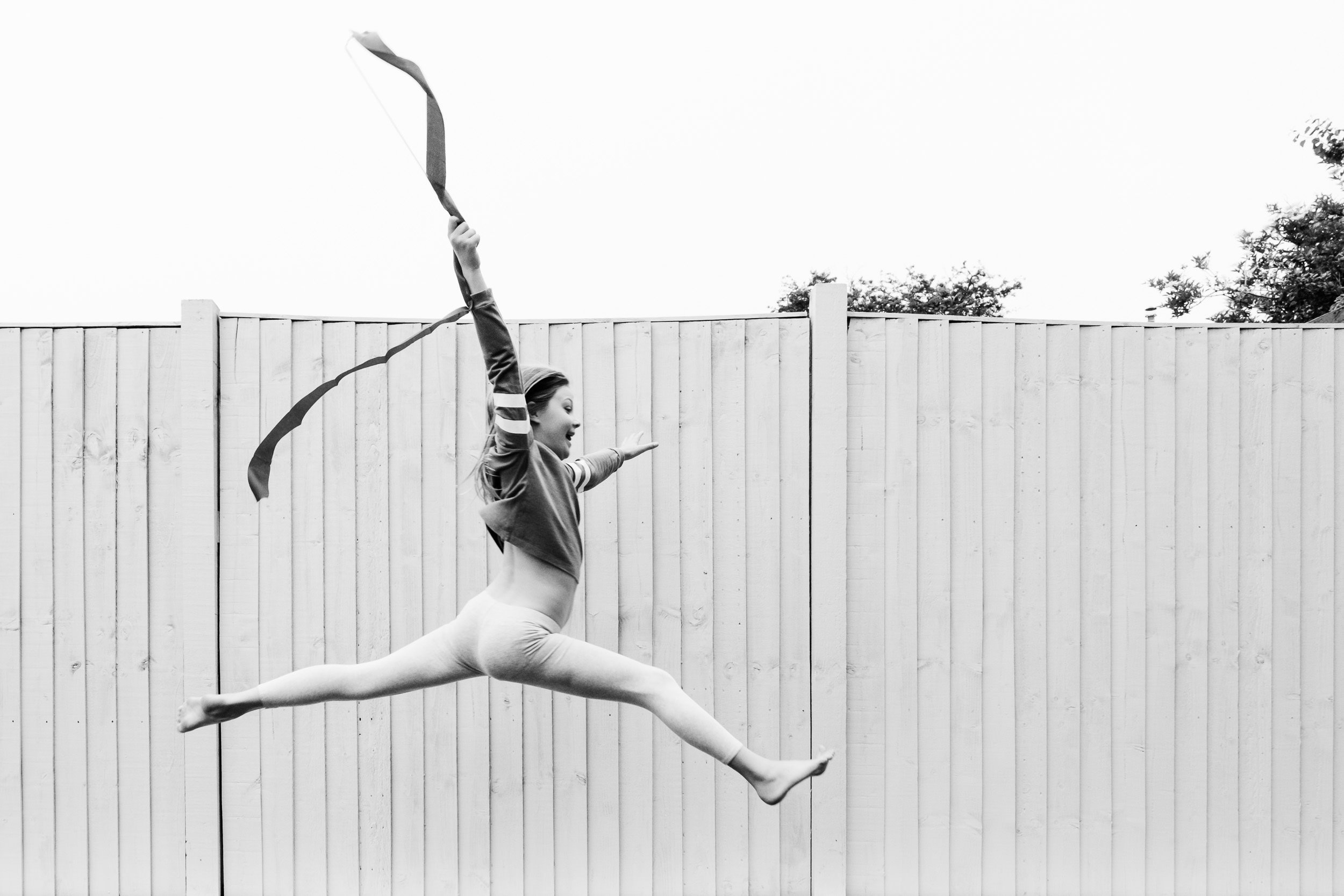 A young leaping, performing a graceful dancer pose with one leg extended behind and arms stretched, against a background of a wooden fence and trees, in black and white.