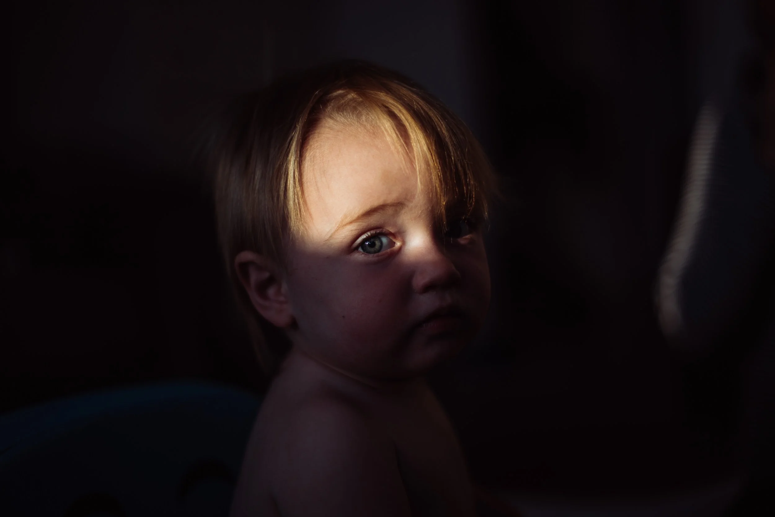 A young child with light-colored hair and blue eyes, looking over their shoulder with a partially shadowed face, against a dark background.
