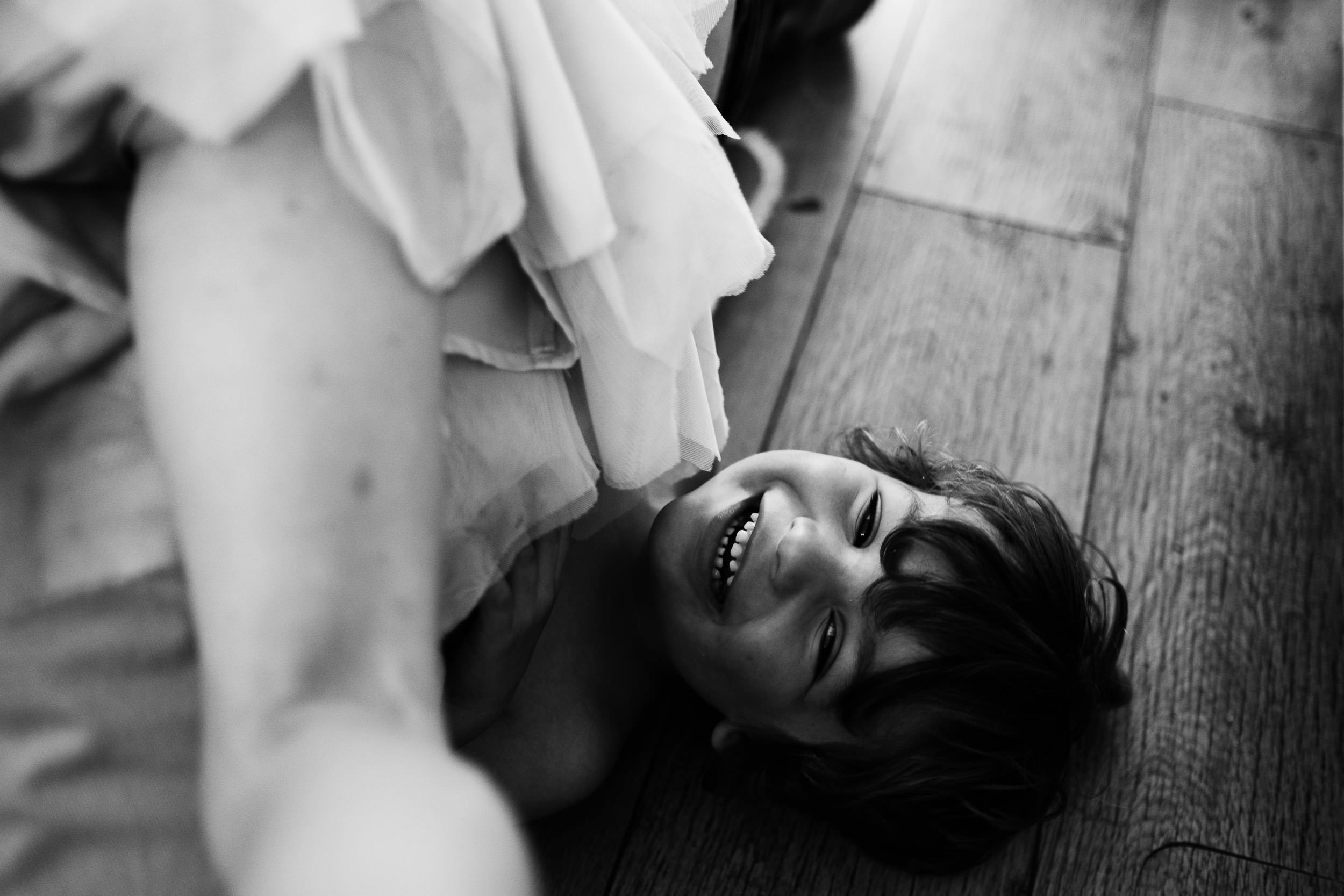 A young boy lying on a wooden floor, laughing, the leg of his sister visible as she lies on top of him.