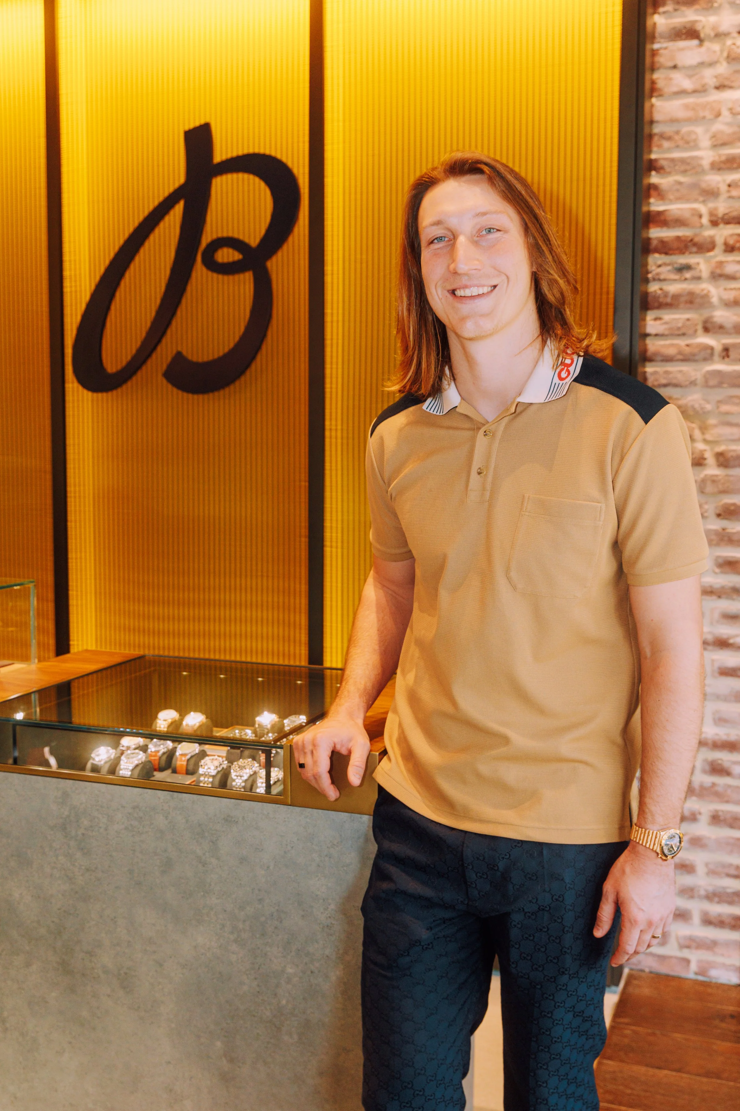 A man with long hair smiling in a jewelry store, wearing a beige polo shirt and patterned black pants, standing next to a display case of watches, with a brick wall and yellow decorative panels in the background.