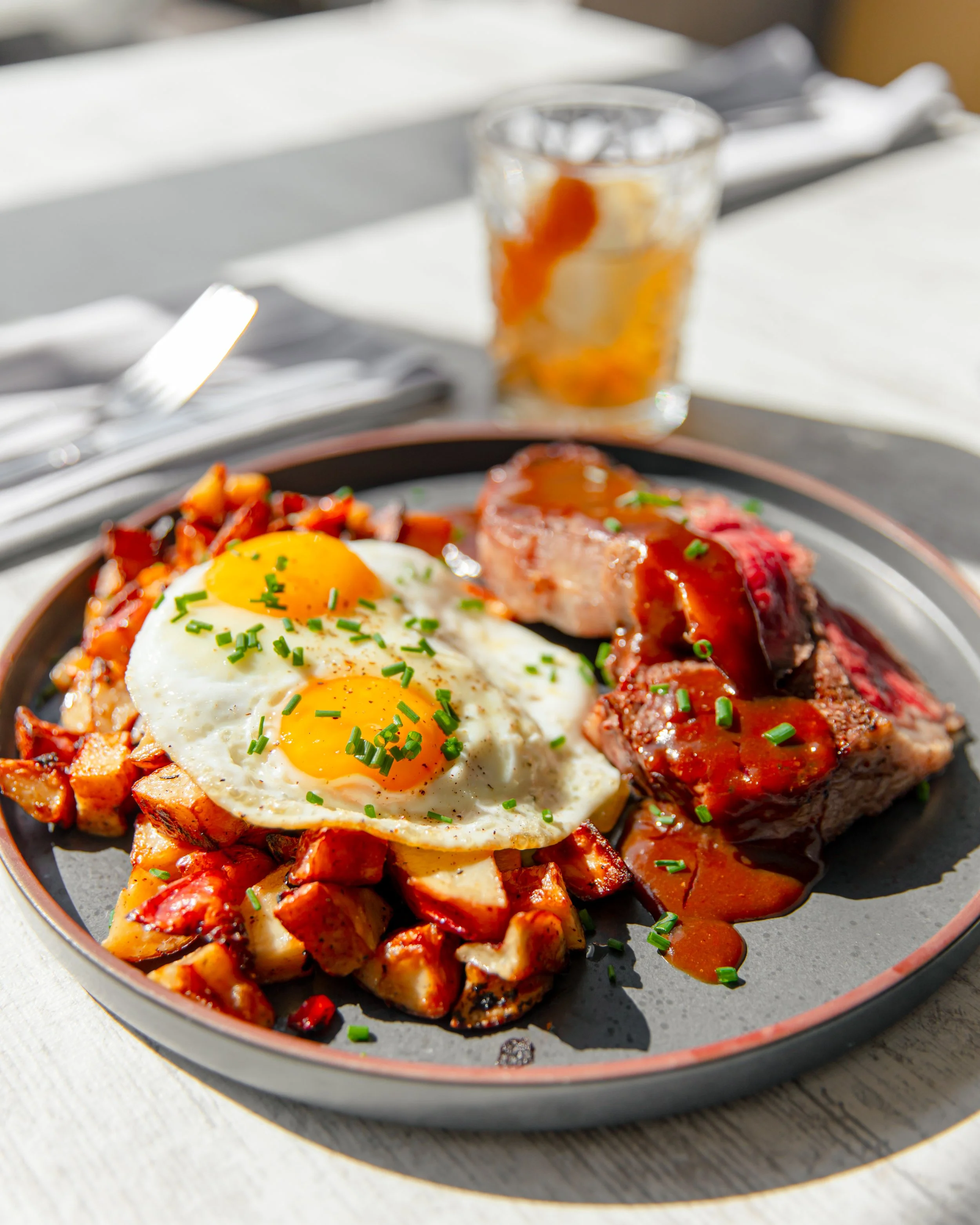 Close-up of a plate with fried eggs, roasted potatoes, sausage, and steak with gravy, garnished with chopped chives. For Coop303.
