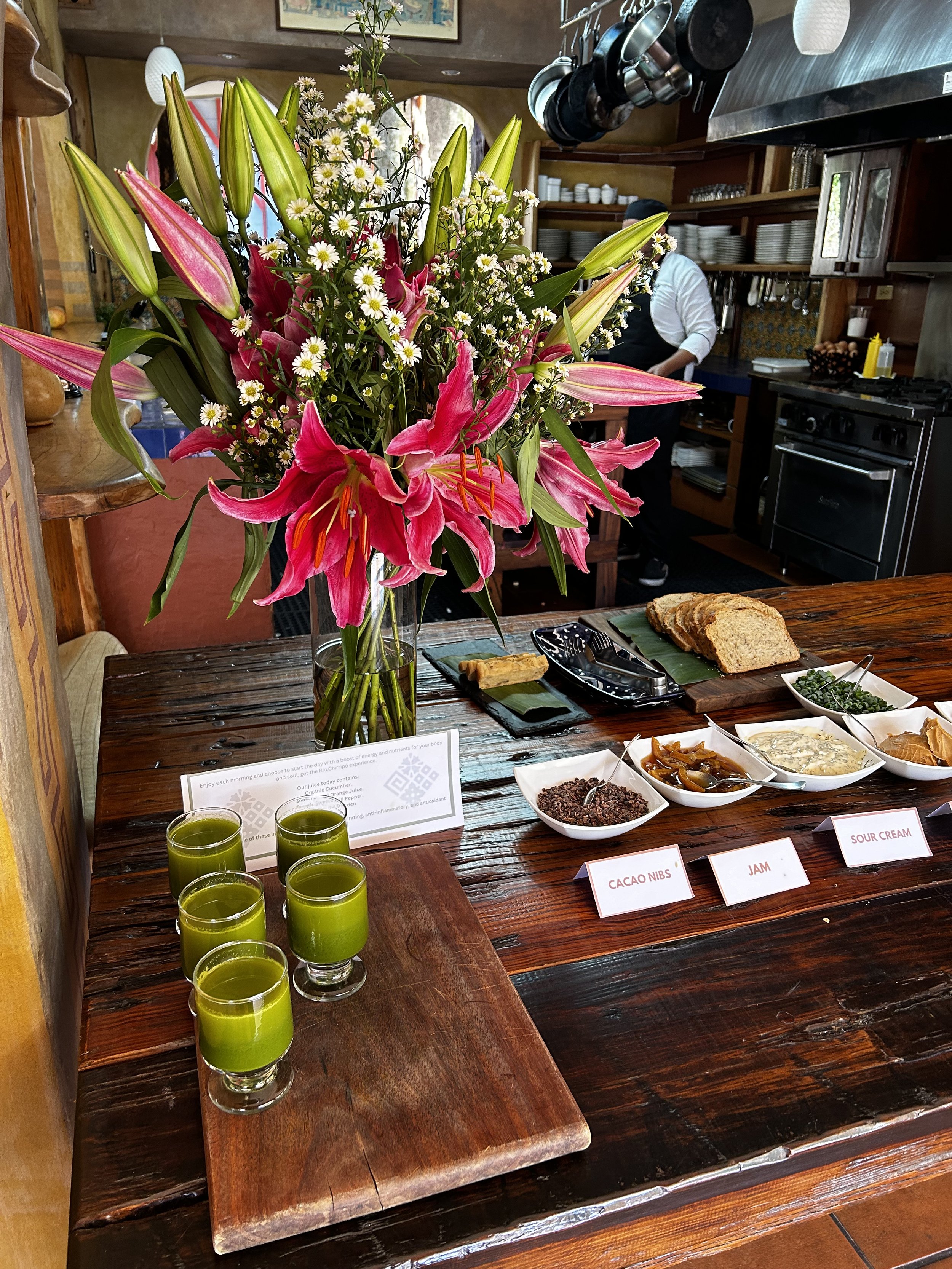 A kitchen island with a large bouquet of pink lilies and white daisies. There are six glasses of green juice on a wooden board, and several small dishes containing cacao nibs, jam, sour cream, and other condiments. A chef is seen in the background.