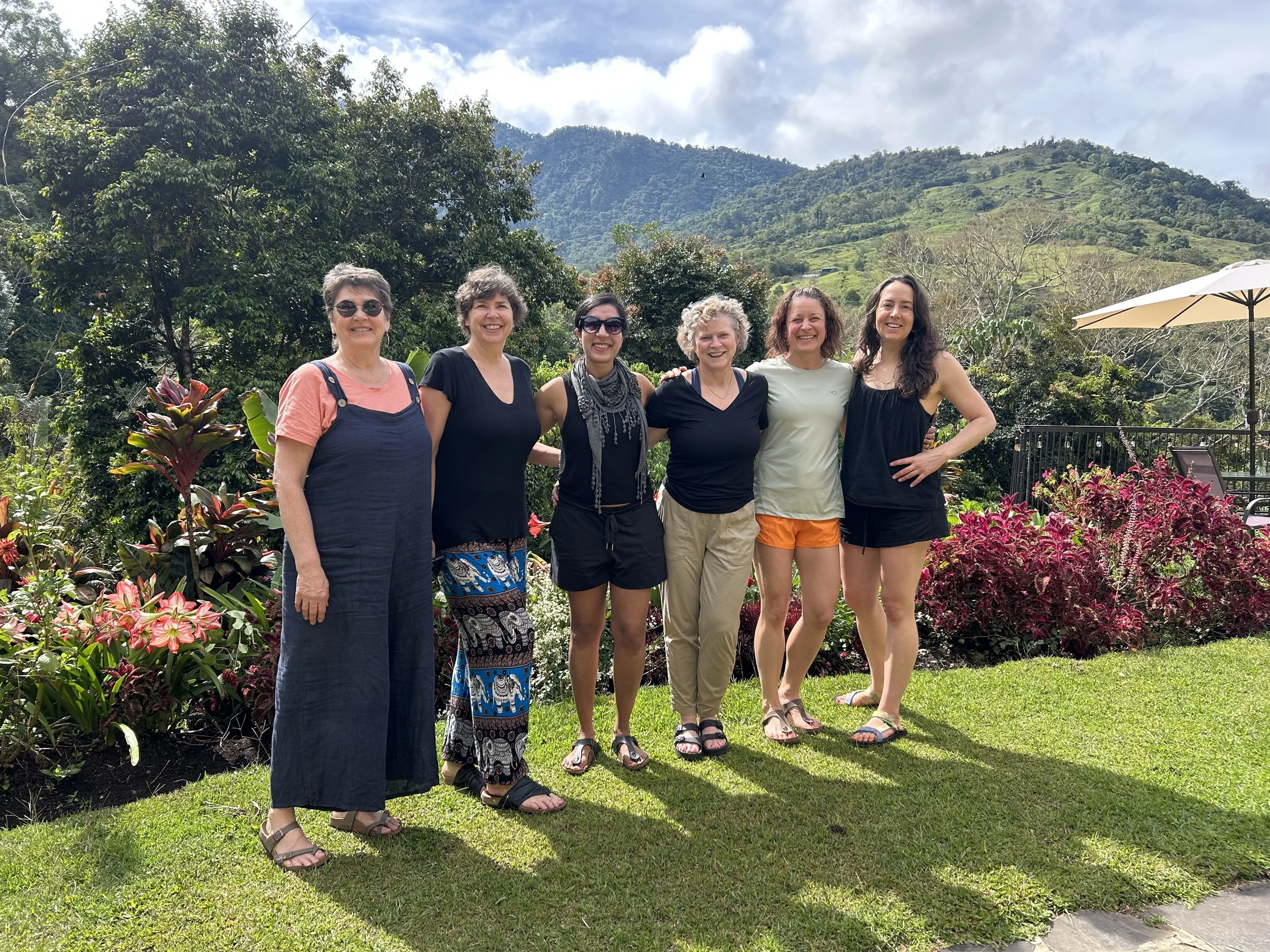 Group of six women posing outdoors in a garden with a scenic mountain backdrop, under a partly cloudy sky.