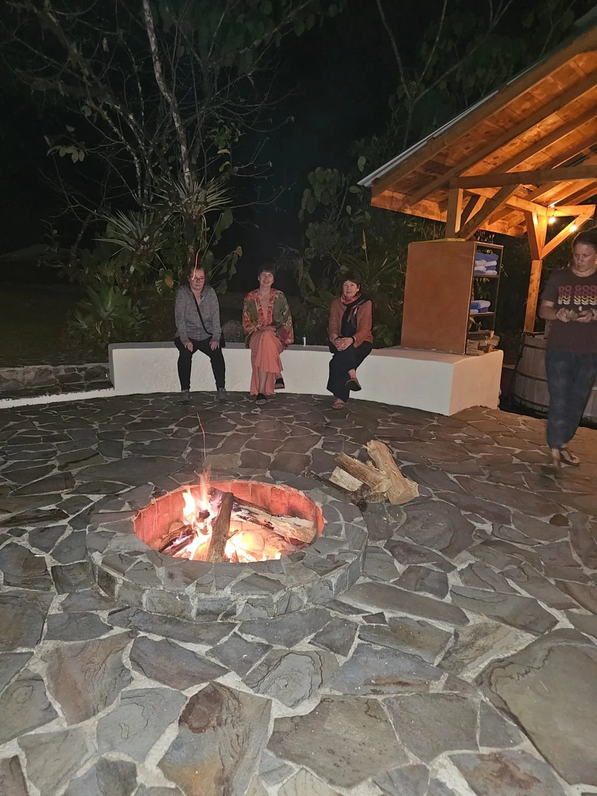 Group of people sitting around a stone fire pit outdoors at night, with a wooden roofed structure in the background.