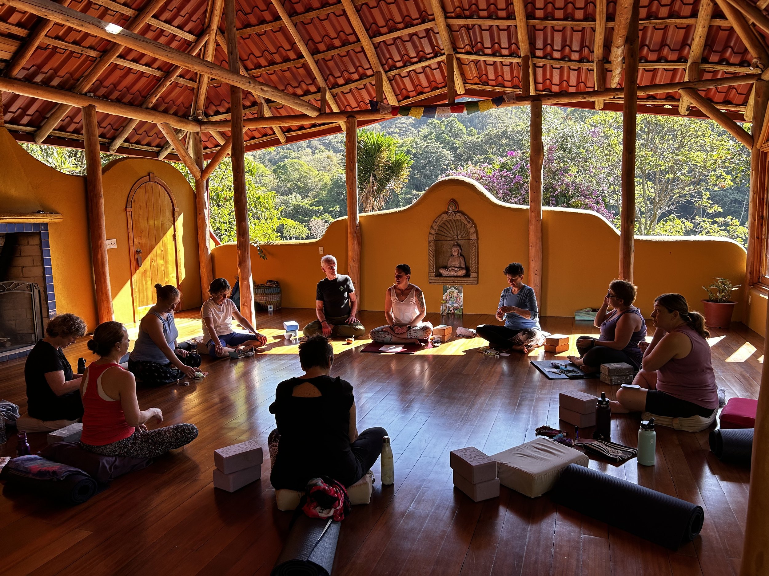 Group of people sitting in a circle inside a wooden meditation room with an open view of nature, sunlight filtering through the roof and walls. Yoga mats, blocks, and cushions are scattered around the floor. A statue is mounted on a wall.