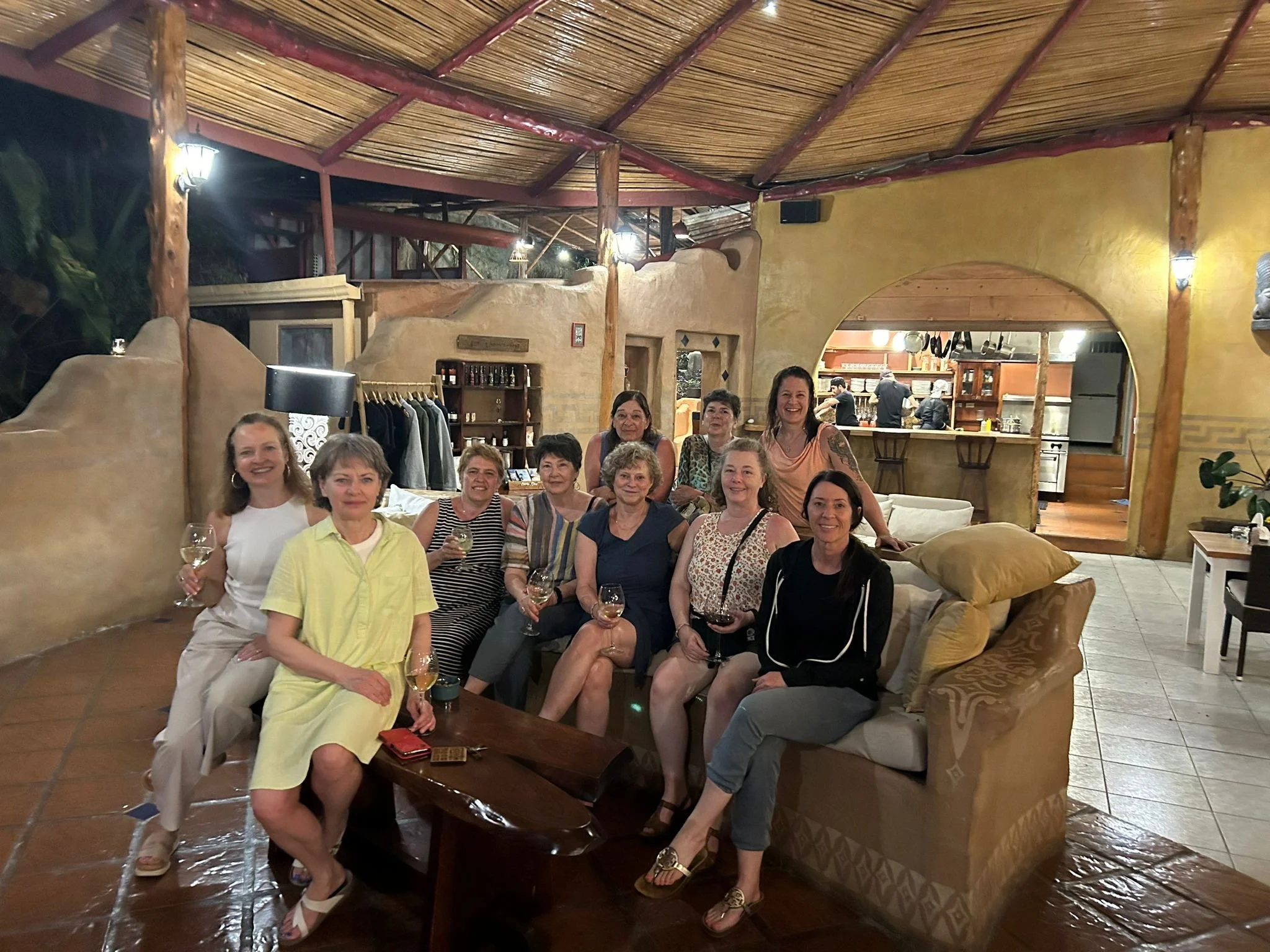 Group of women sitting and standing in a cozy, rustic indoor setting, holding wine glasses, under a bamboo roof with warm lighting.