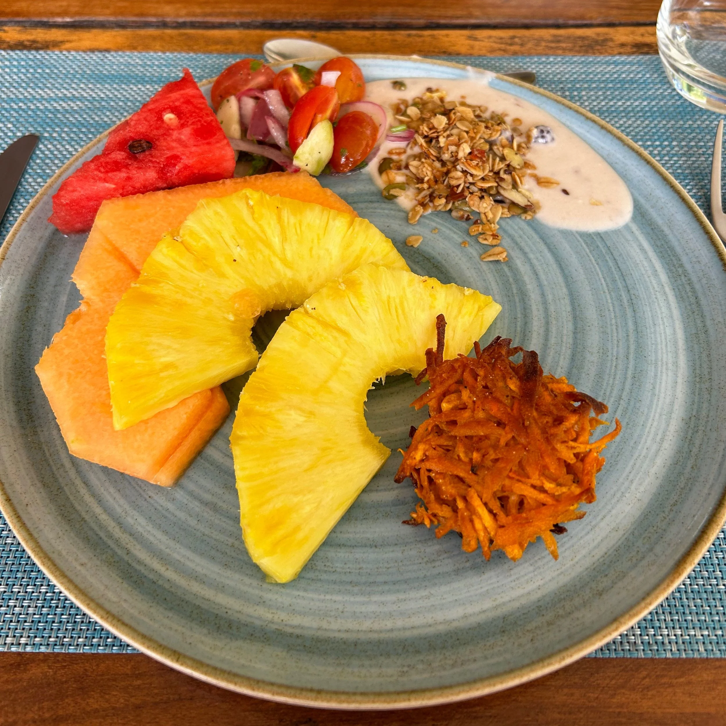 Plate with sliced pineapple, watermelon, cantaloupe, a small salad with tomatoes and onions, granola with yogurt, and a hash brown on a blue plate.