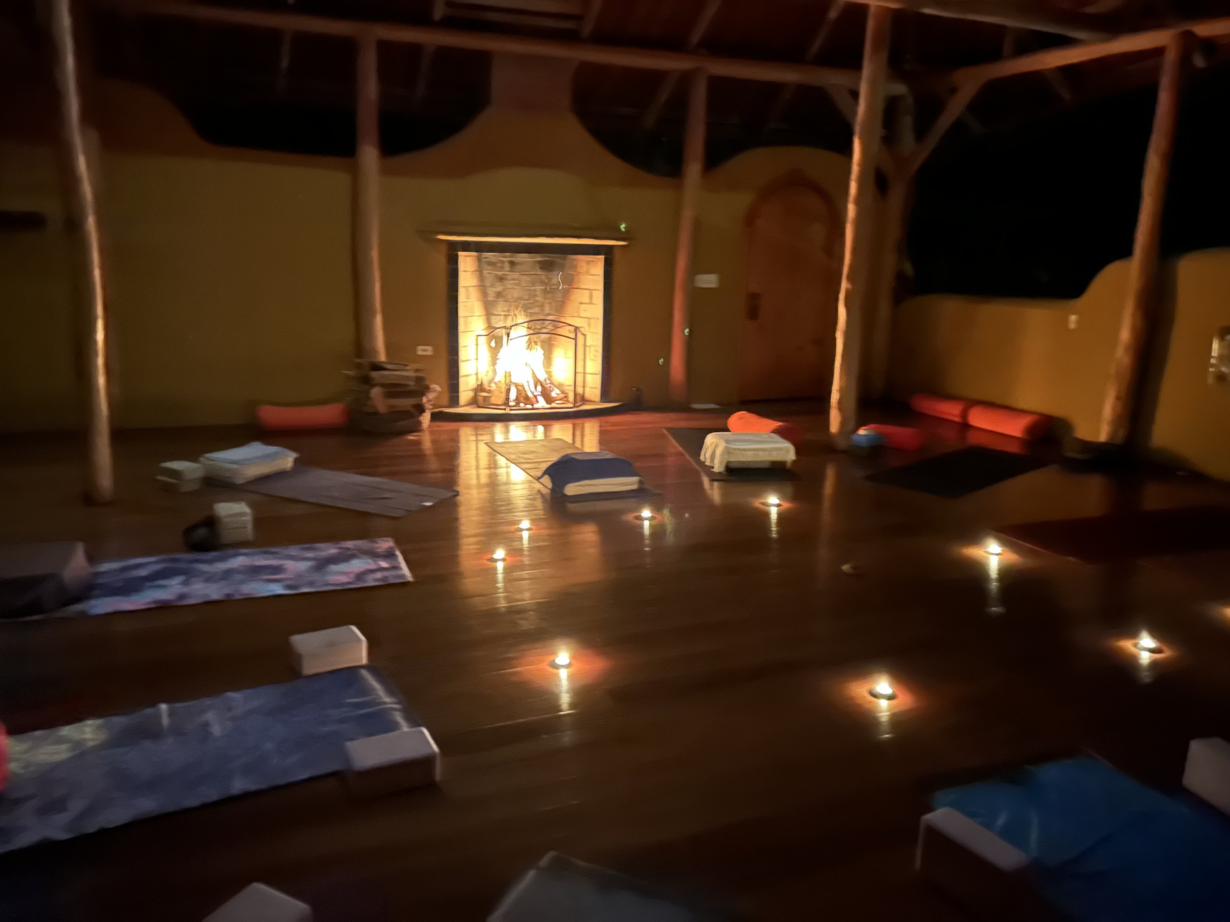 Yoga studio with wooden floor and ambient lighting, featuring yoga mats, bolsters, blocks, blankets, and a lit fireplace, surrounded by candles for a cozy and tranquil atmosphere.