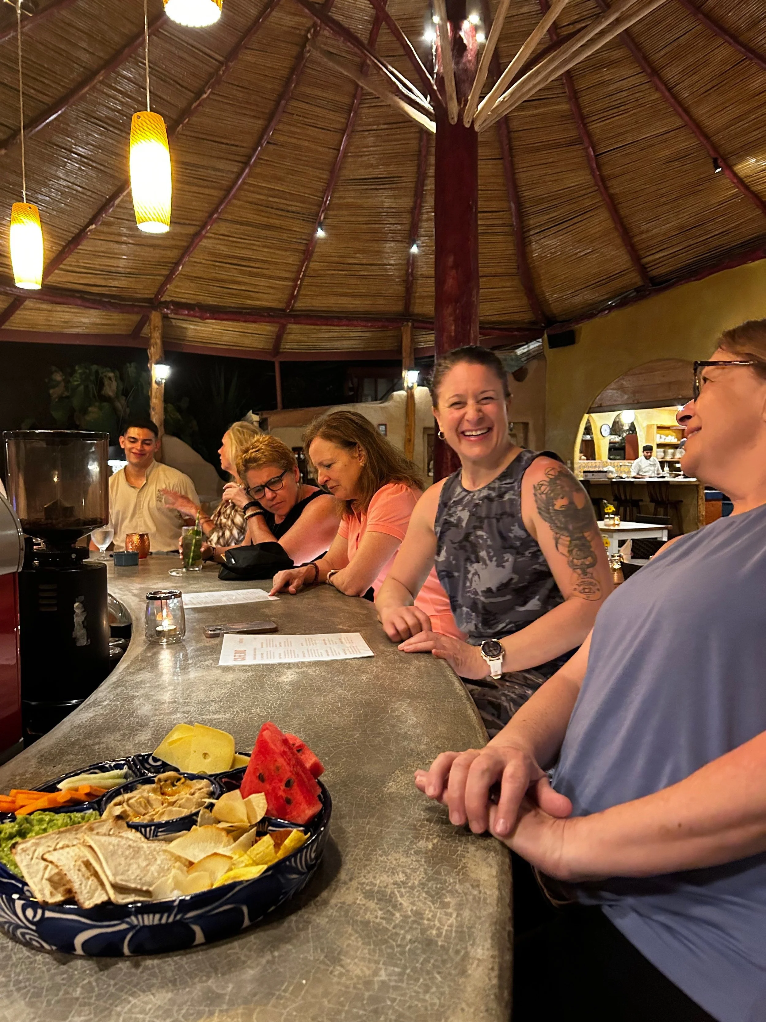 Group of people chatting and laughing inside a thatched-roof bar, with a platter of various snacks including watermelon and chips on the counter.