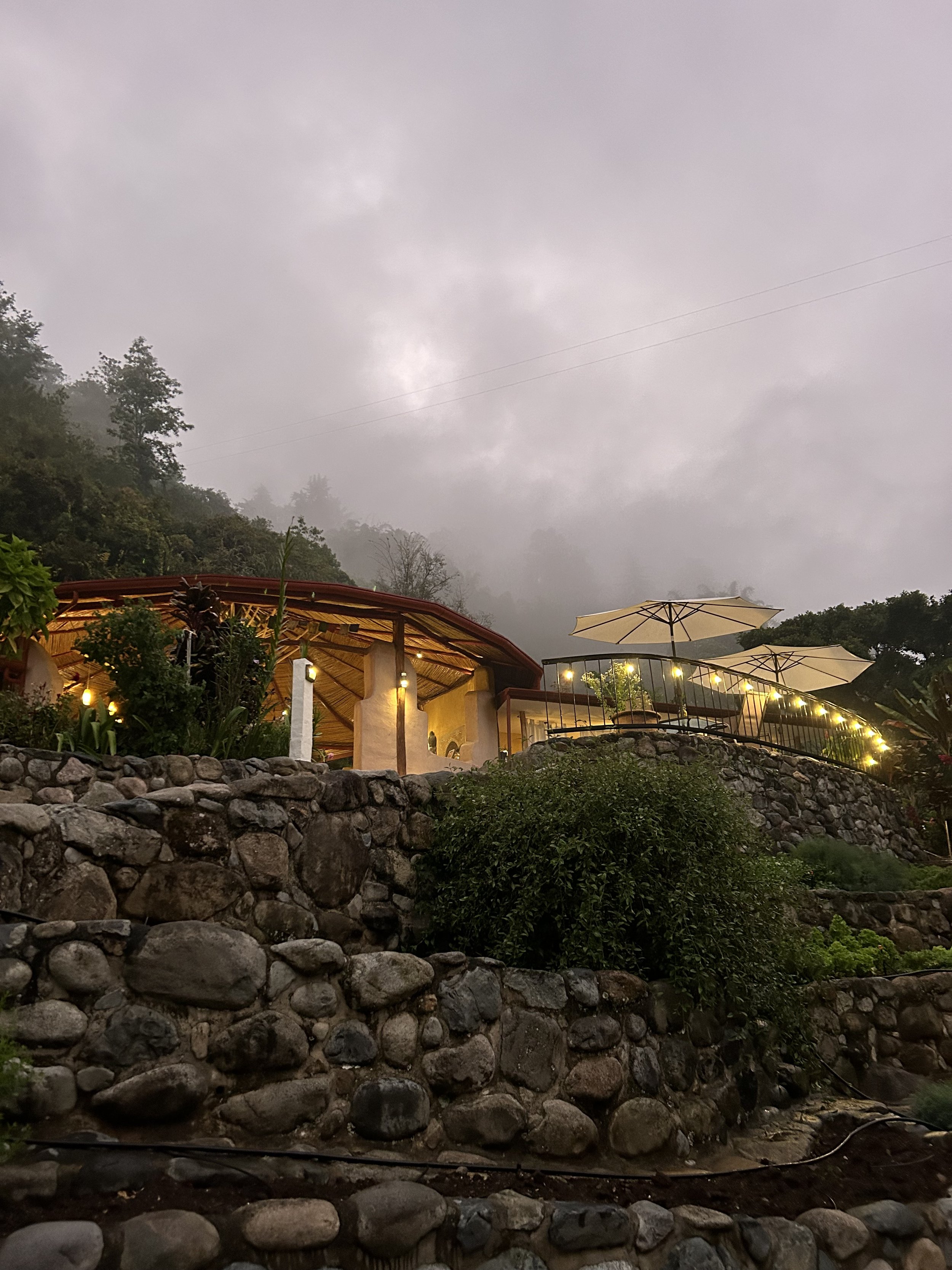 A hillside terrace with a stone wall and a house surrounded by trees. The sky is overcast with gray clouds, and the setting appears to have outdoor umbrellas and chairs.