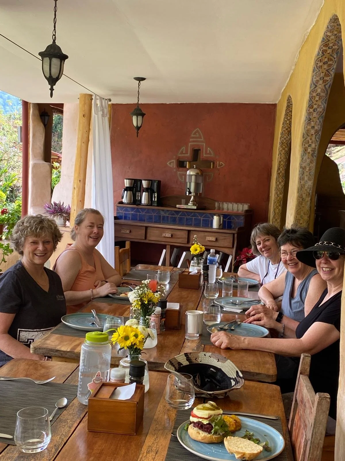 A group of six women sitting around a wooden dining table, enjoying a meal in a rustic setting. The table is set with plates, utensils, and glasses. Flowers and decorative items adorn the table. The background features a decorative wall with a coffee station. Natural light filters in, creating a cozy atmosphere.