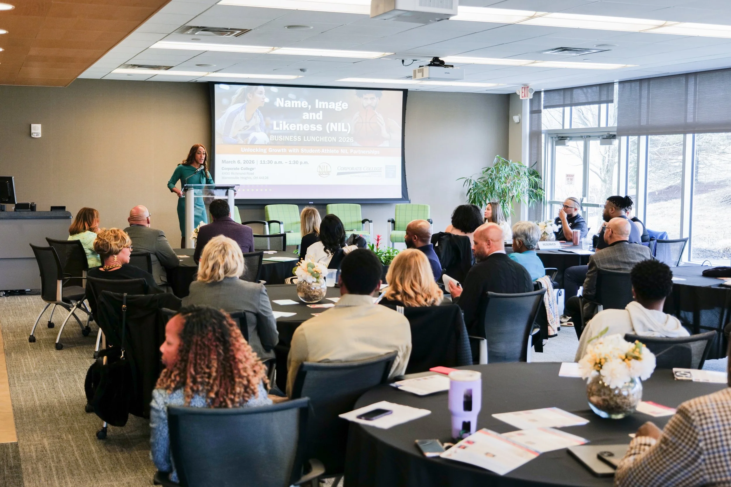 A conference room filled with people sitting at round tables listening to a woman speak at a podium. The room has large windows, plants, and a large screen displaying a presentation titled "Name, Image and Likeness (NIL) BUSINESS LUNCHEON 2026".