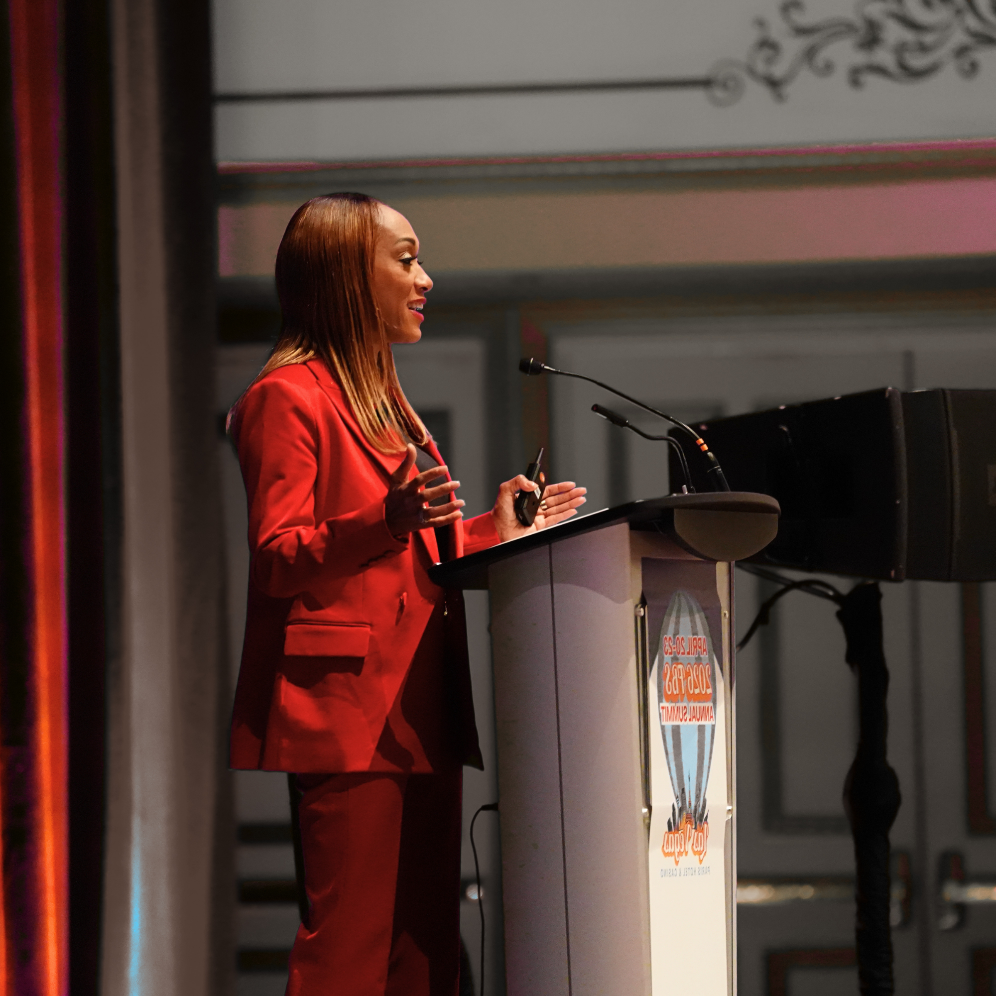A woman with long red hair wearing a red blazer speaking at a podium with a microphone. She is standing in front of a large screen displaying a presentation slide at an indoor event.