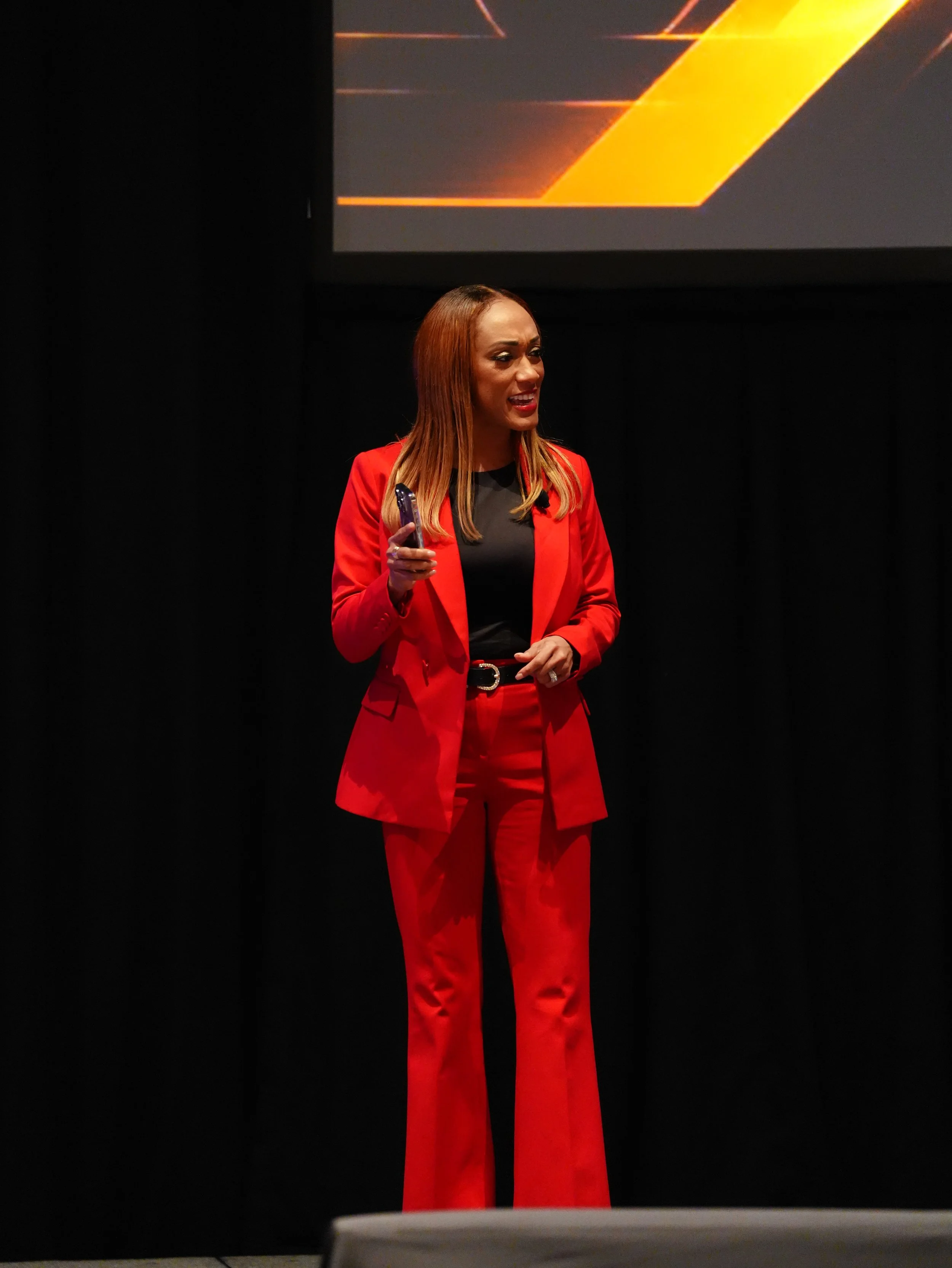 A woman with long, reddish-brown hair, wearing a red suit with a black top, standing on stage, speaking into a microphone, with a dark background and a dark screen displaying an orange and black graphic in the top right corner.