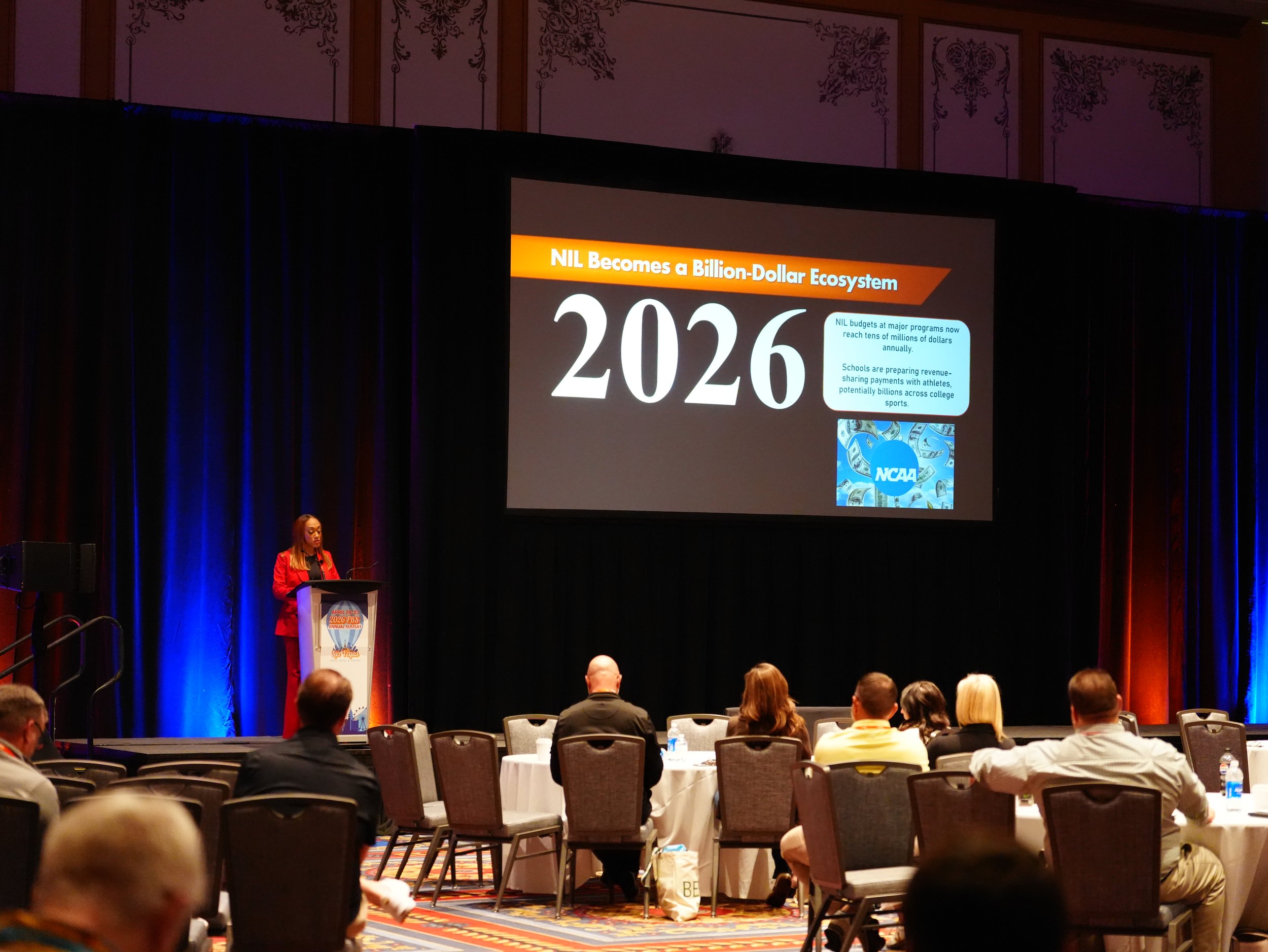 At a conference with a large screen displaying the title 'NIL Becomes a Billion-Dollar Ecosystem 2026' and NCAA logo, a woman is speaking at a podium with seated audience members in the foreground.