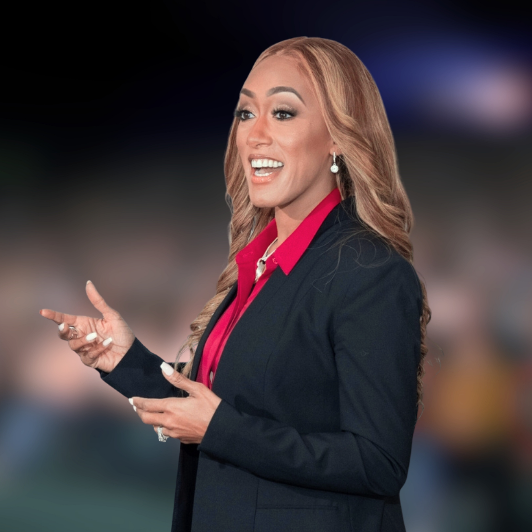 A woman with long wavy reddish-blonde hair, dressed in a black blazer and red blouse, speaking and gesturing with her hands, standing against a blurred dark background.