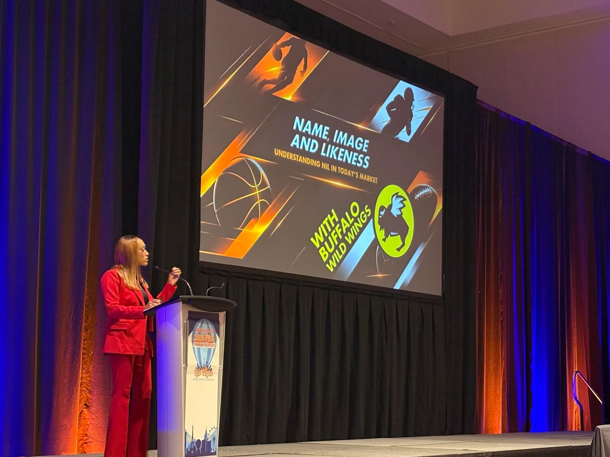 A woman dressed in a red suit speaking at a podium during a conference or summit in a large room with dark curtains. A large screen behind her displays a slide with a football player graphic, the text 'NAME, IMAGE AND LIKENESS', and logos including 'The Buffalo Wings Wild Wings' and an illustration of a hot air balloon.