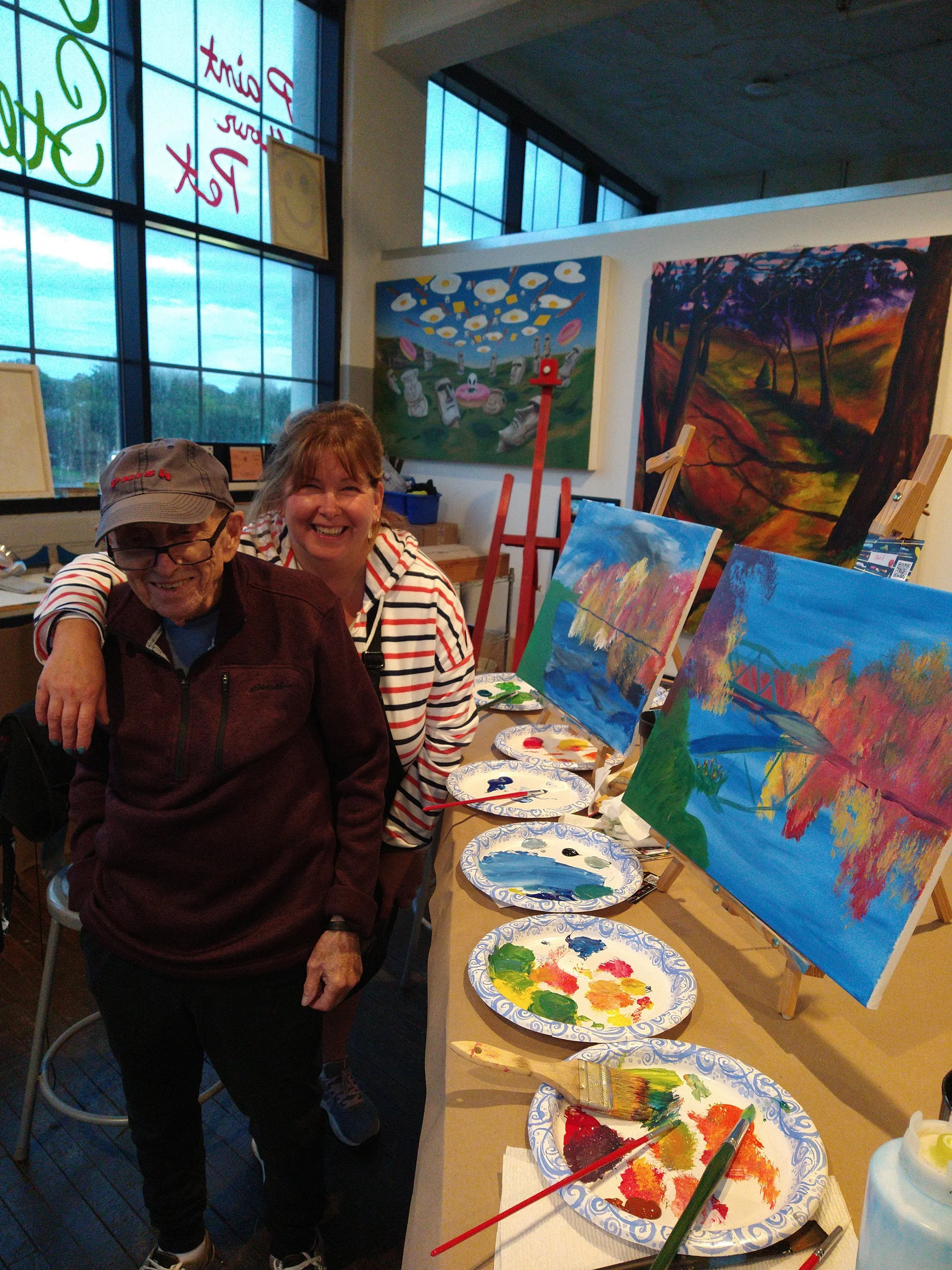 Two smiling people in an art studio stand near painted canvases on easels. The table in front of them has paper plates with colorful paint and brushes. The background shows large windows and two paintings on the wall.