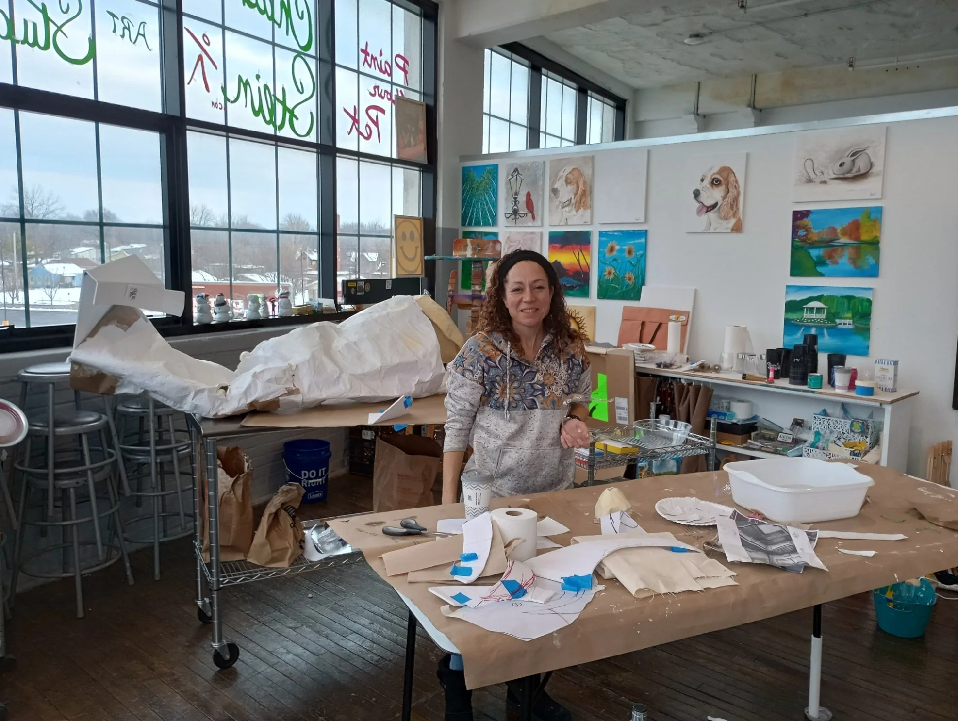 A woman standing in an art studio with various paintings on the walls, art supplies on the tables, and large windows in the background. The studio is filled with creative materials and art projects in progress.