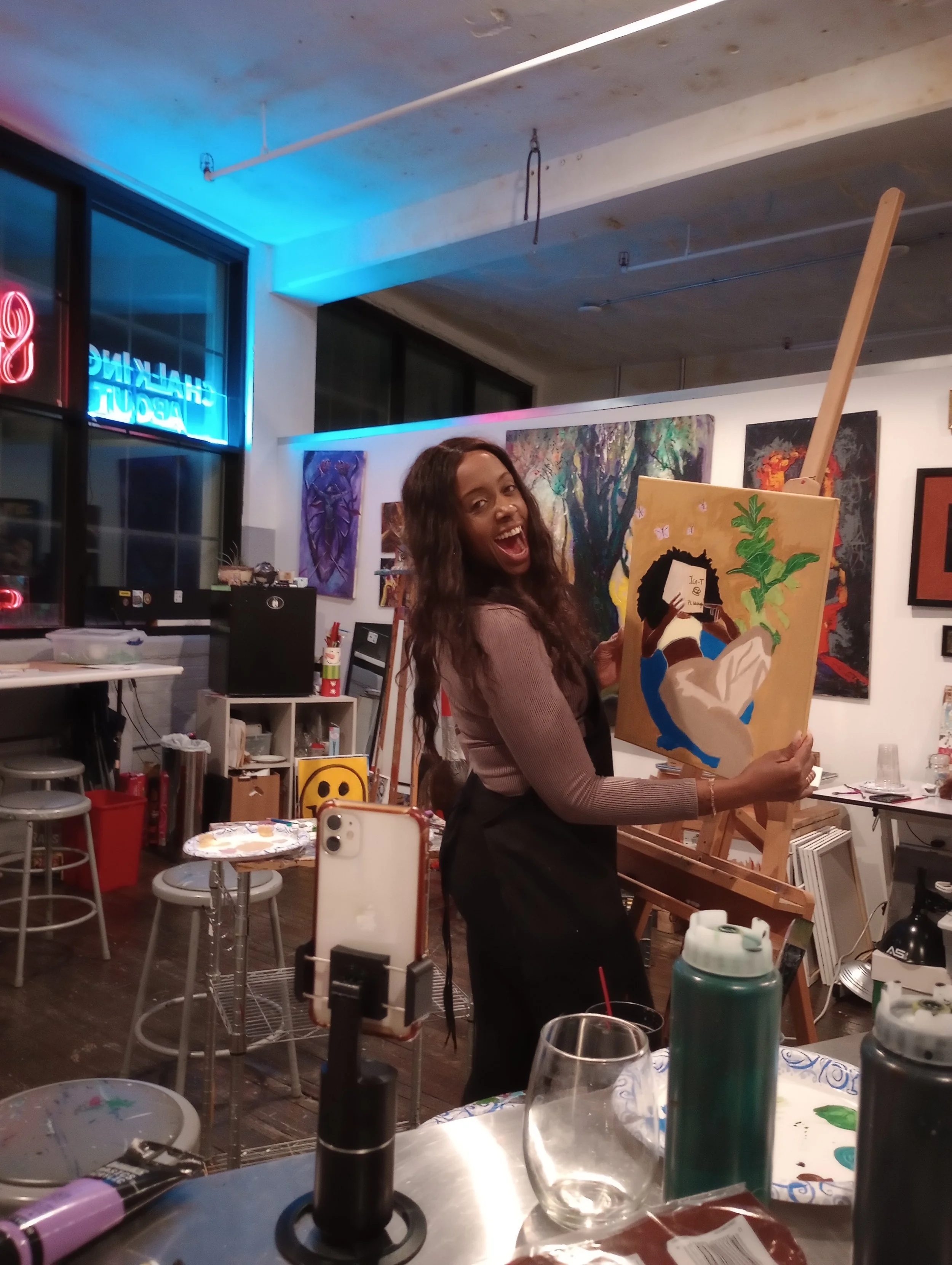 A smiling person in an art studio holding a canvas with a painting. They are wearing a brown top and black apron. The studio has paintings on the walls and art supplies scattered around. The environment is well-lit with neon and fluorescent lights.