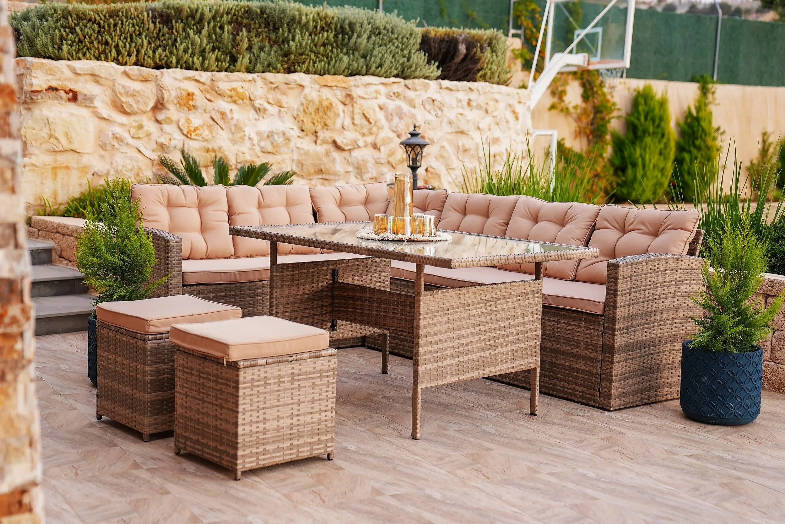 Outdoor patio with wicker sectional sofa with cushions, a glass-top table, small wicker ottomans with cushions, potted plants, and a stone wall with lush greenery in the background.