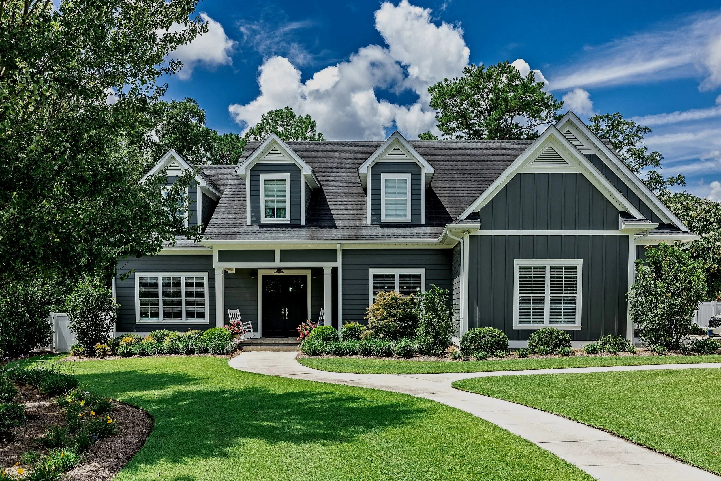 A large, two-story house with dark blue exterior siding, white trim, and multiple dormer windows on the roof. The front yard has a neatly manicured lawn, a curved concrete walkway, and various bushes and small trees. There are two white rocking chairs on the porch, and the sky above is blue with some white clouds.