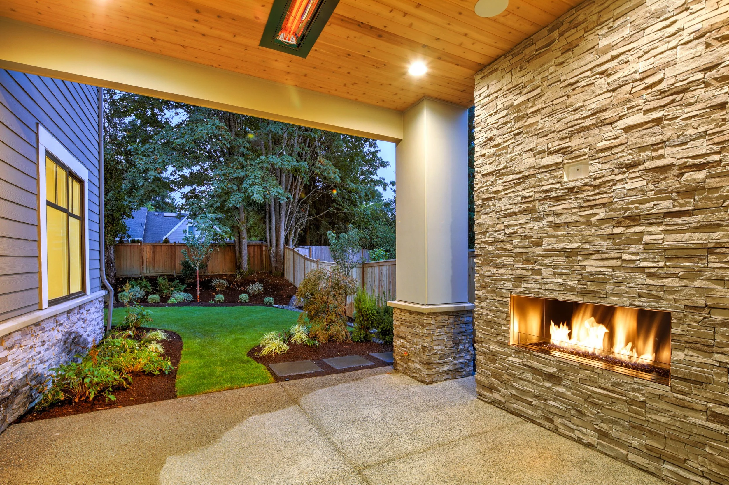 Backyard gas fireplace with stone veneer finish, matching pillars, wood privacy fence, and landscaped outdoor living space in Delaware by Studio G Outdoor Living