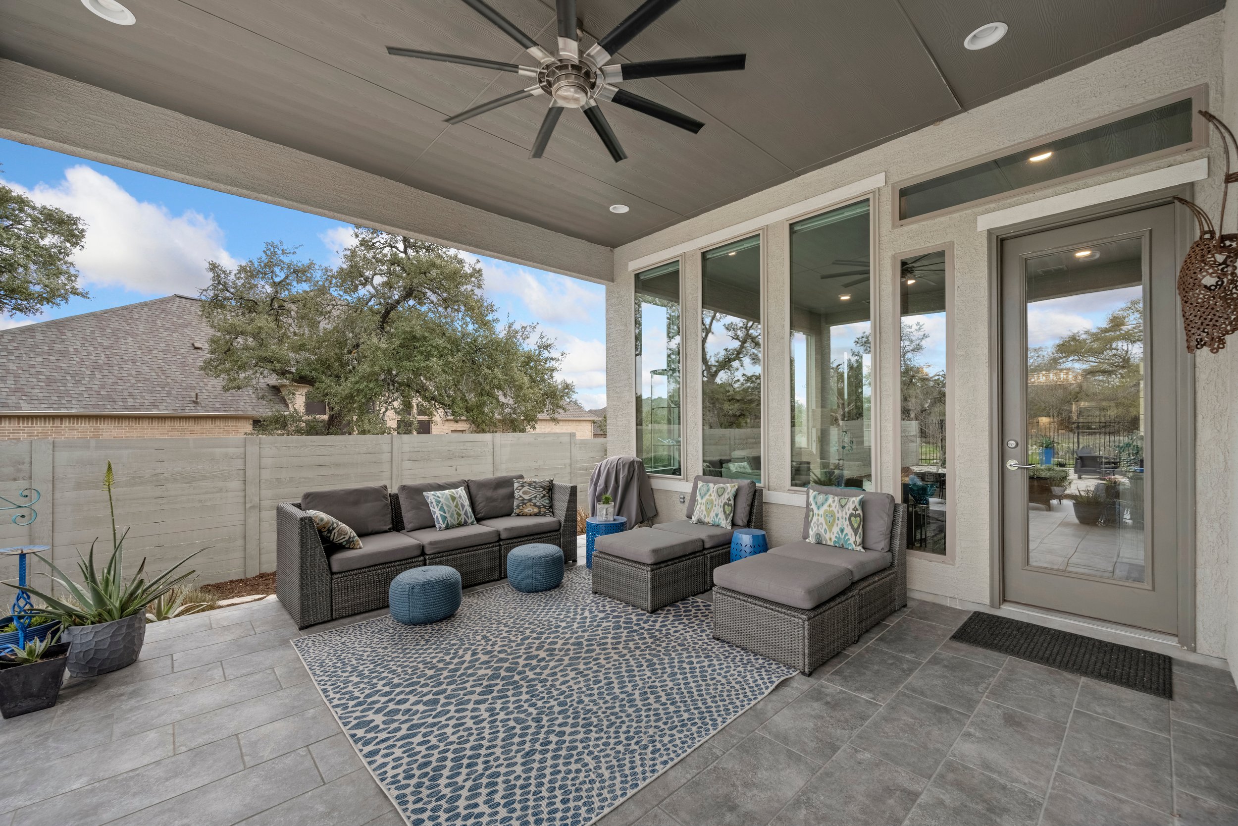Covered outdoor patio with gray wicker sofa and chairs, blue and patterned pillows, small blue stools, potted plants, and a patterned outdoor rug, overlooking a backyard with trees and a wooden fence.