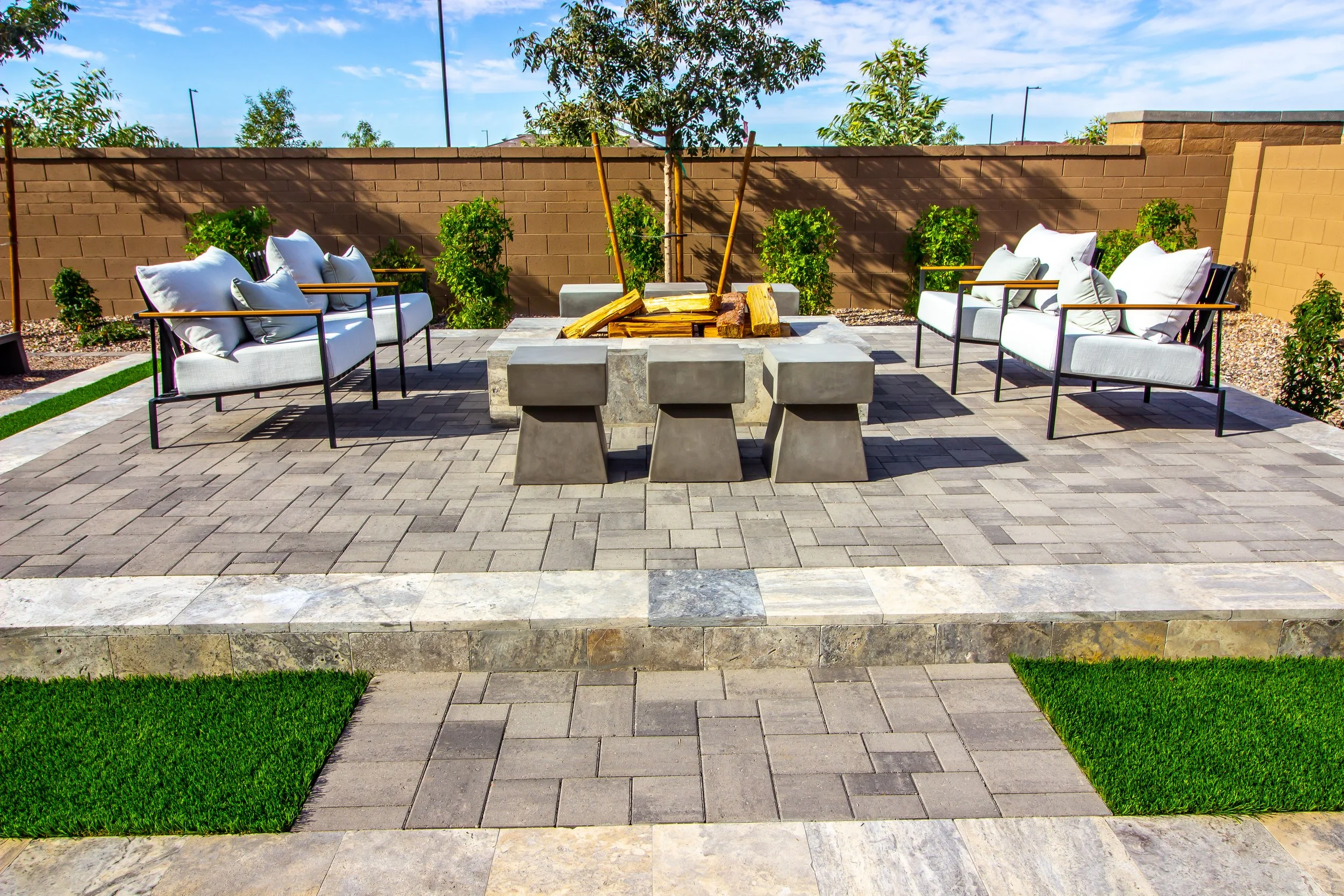 Outdoor patio with two white sofas, a central firepit filled with wood, a brick wall with trees, and a blue sky.