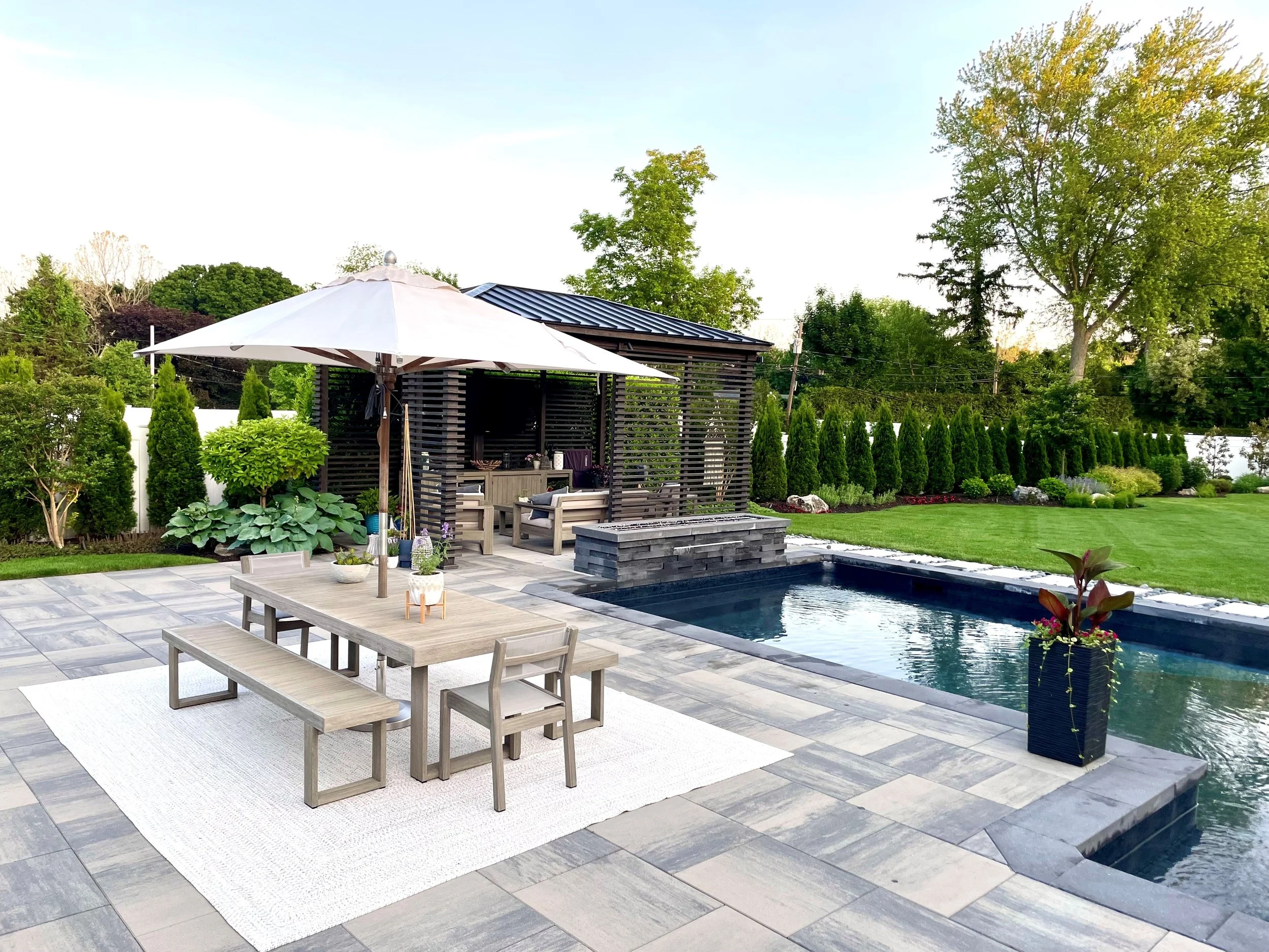 Backyard with a patio, outdoor dining table, pool, lush green lawn, and landscaped garden with trees and bushes.